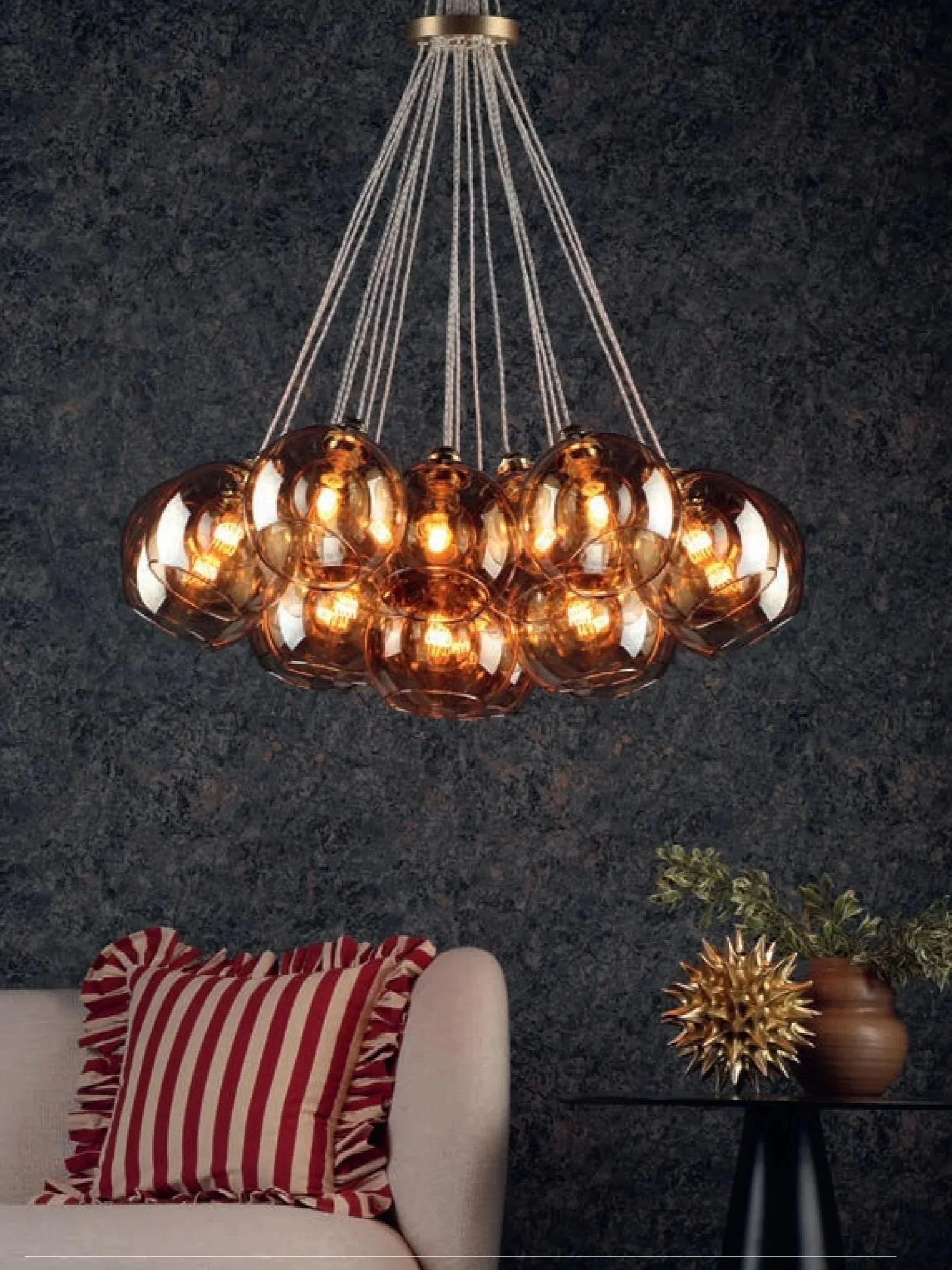 A modern interior living room with a beige sofa, red striped pillow, dark textured wall, gold and glass chandelier hanging from ceiling, and a black side table with a gold spiked decorative object and a brown pot with green plants.