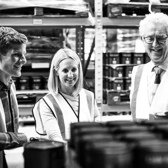 Three people smiling and talking in a warehouse or storage facility with shelves and boxes in the background.