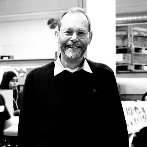 A smiling man with glasses, a beard, and a mustache, wearing a dark sweater over a collared shirt, standing in a room with shelves and other people in the background.
