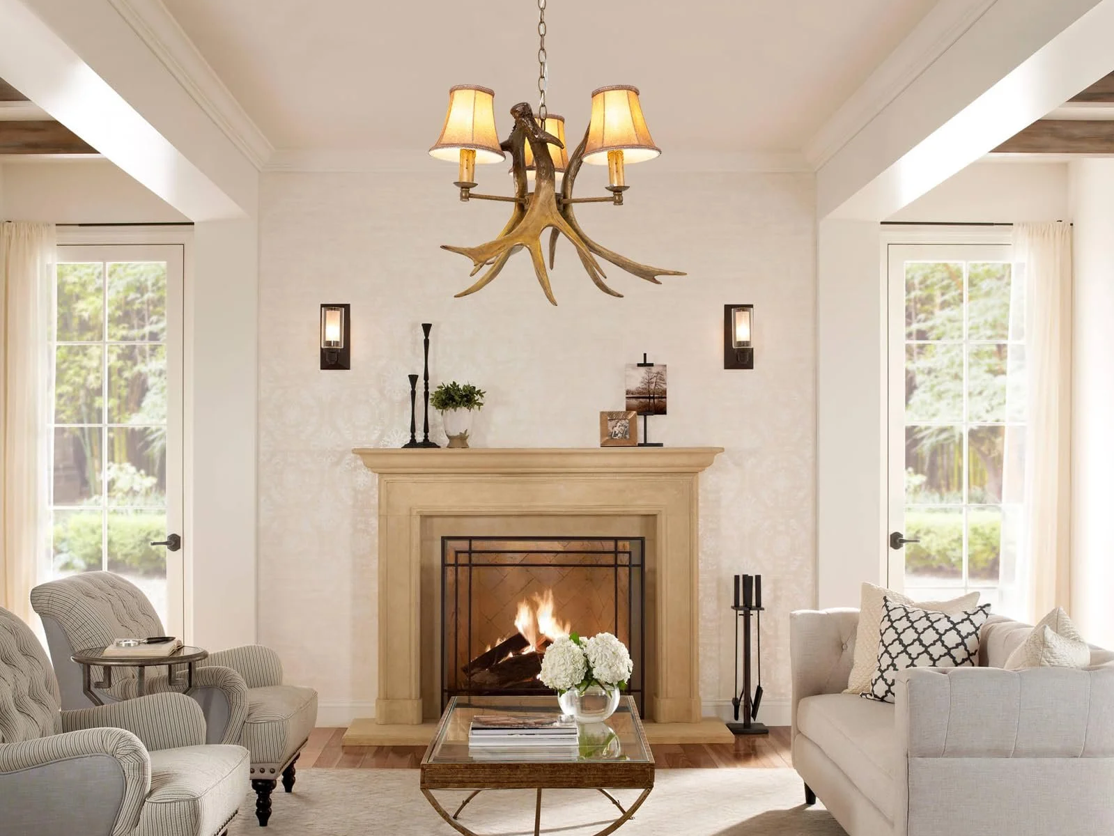 Living room with a fireplace, beige mantel, and antler chandelier. Two beige armchairs and a white sofa with black and white pillows. Coffee table with flowers, candlesticks, and books. Windows with curtains and decorative items on the mantel.