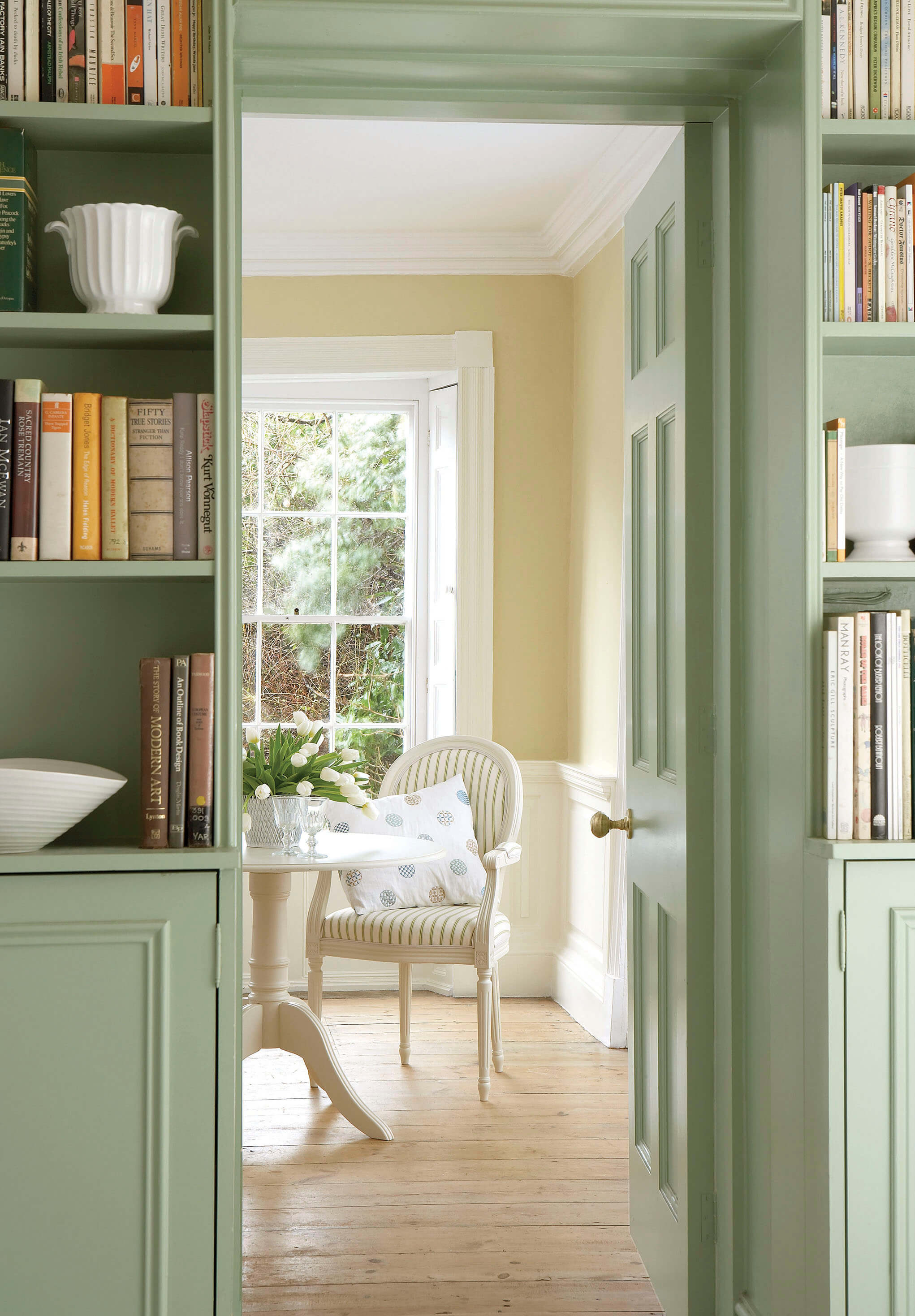 View through a green doorway into a bright room with a window, a round table with flowers, a striped armchair, and bookshelf.