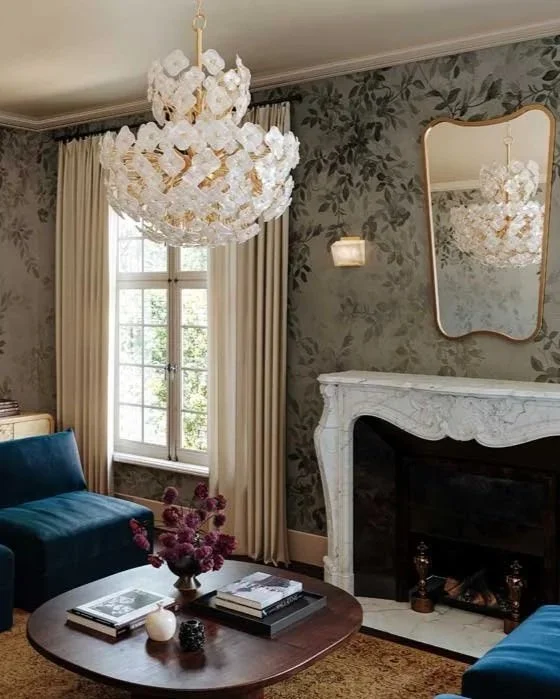 Living room with floral wallpaper, white curtains, a white ornate fireplace, and a large mirror above it. A blue sofa, a round wooden coffee table with books and a flower arrangement, and a chandelier hang from the ceiling.
