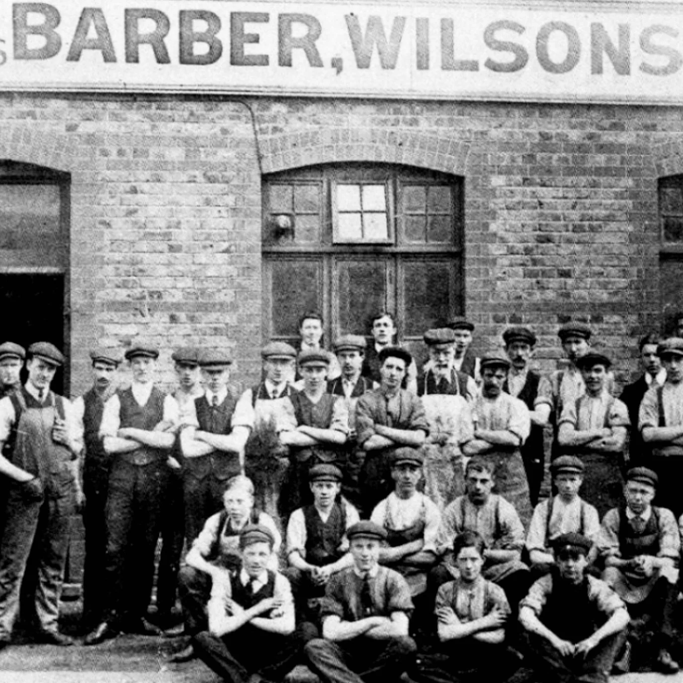 Vintage black and white photograph of a group of young boys and men standing in front of a brick building with a sign that reads "Barber, Wilson." The group appears to be employees or students of the barber shop.