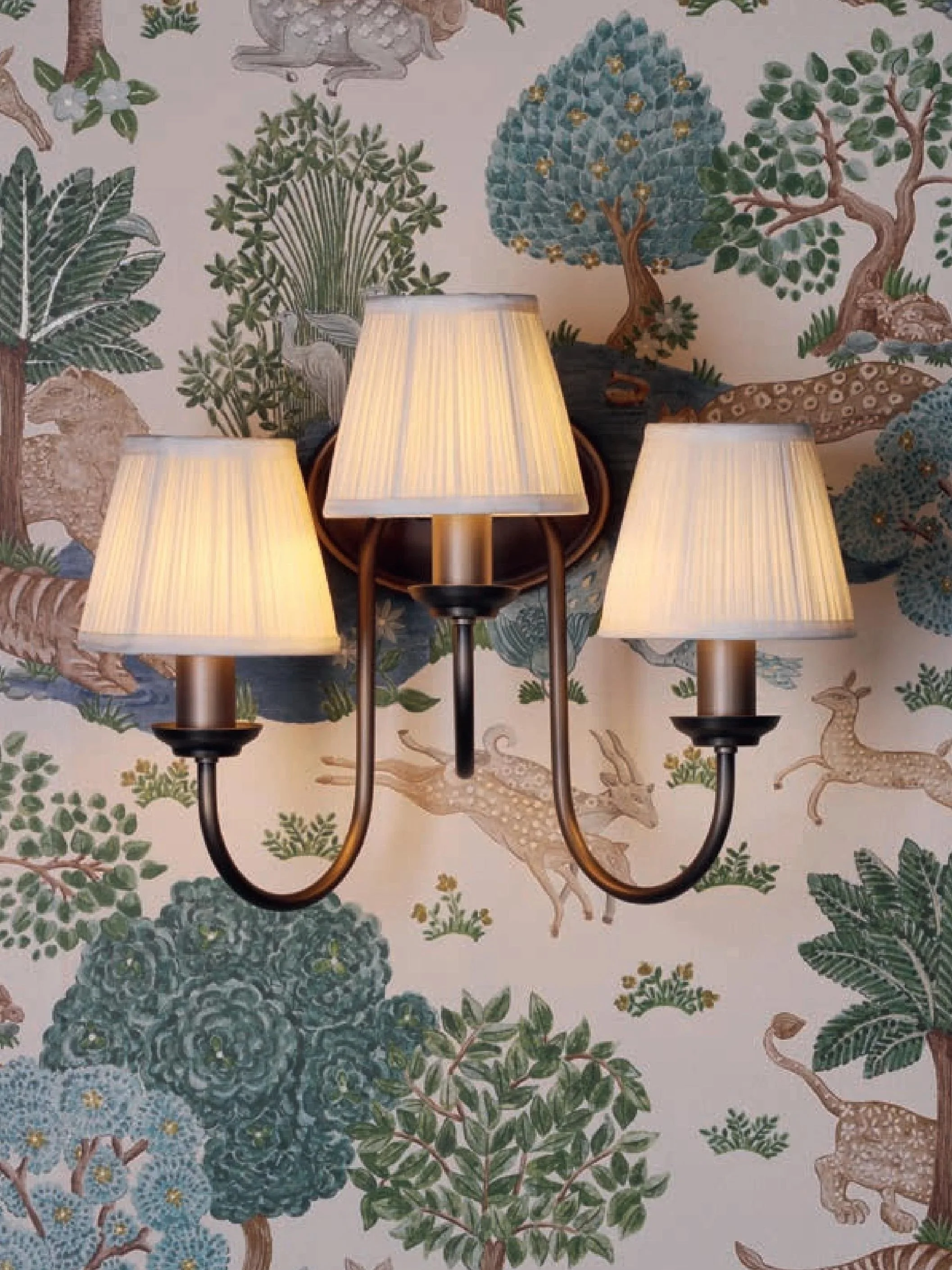 A wall-mounted sconce with three beige fabric lampshades is illuminated and mounted on a wallpaper decorated with various woodland animals and trees, including deer, rabbits, and bears, in a nature-themed pattern.