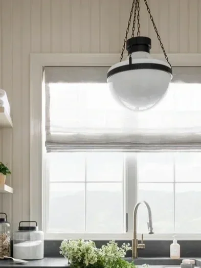 Kitchen window with white blinds, hanging light fixture, and a sink with a faucet, soap dispenser, and plants nearby.