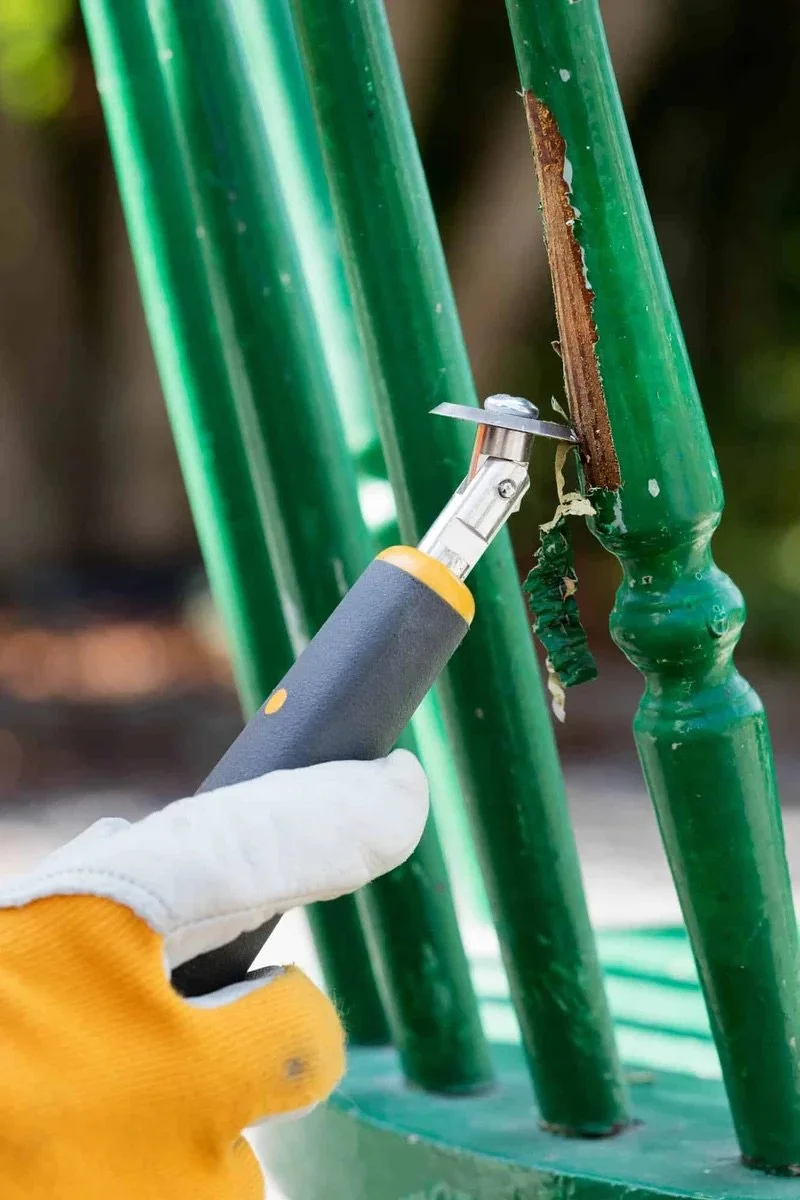 A person wearing a white glove and yellow sleeve is using a utility knife to cut a strip of green paint from a metal railing.