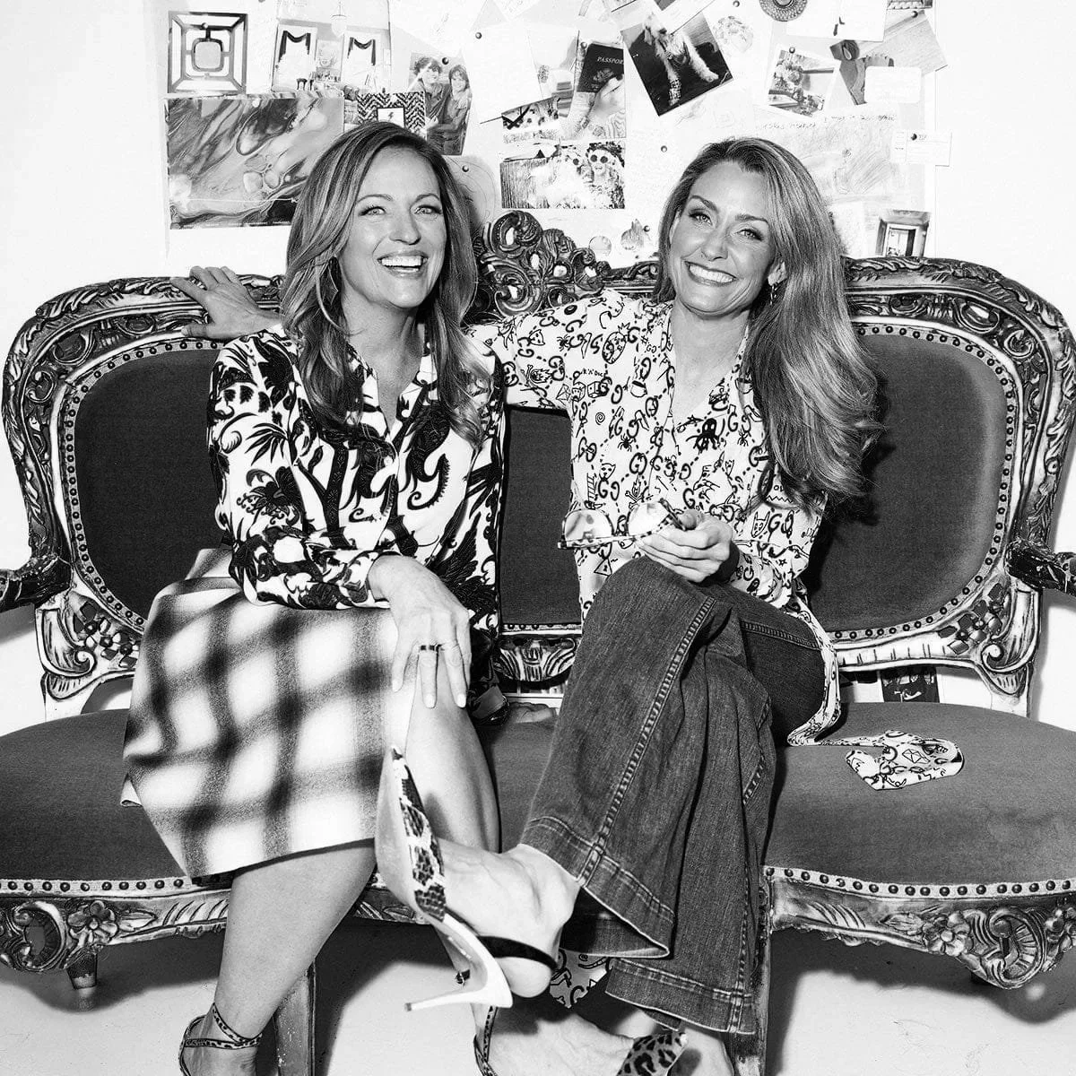 Two women sitting on an ornate vintage sofa, smiling and posing for the camera, with pictures and posters on the wall behind them in a room.