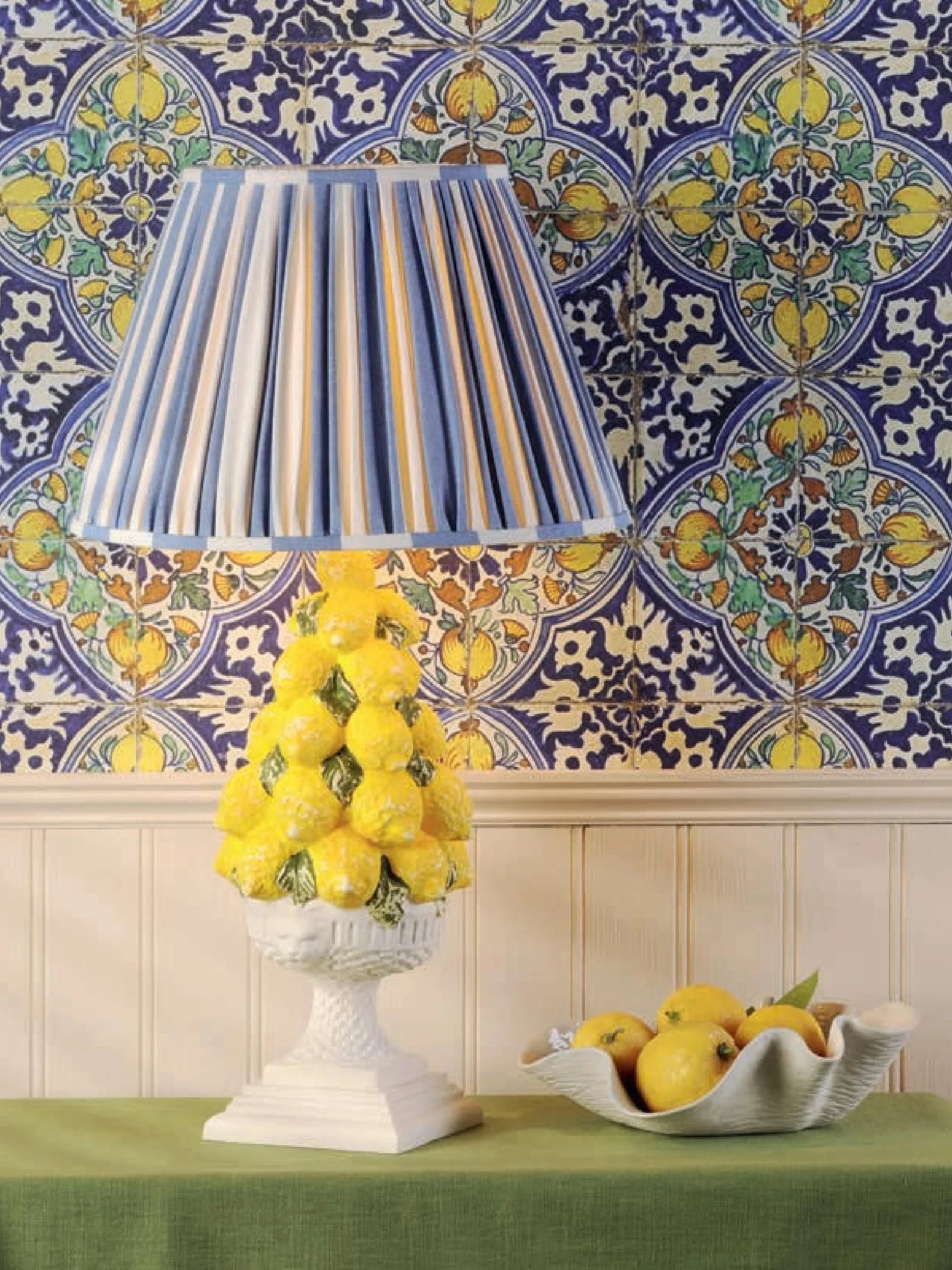 Table with a green cloth holding a decorative lamp shaped like a bunch of lemons and a fruit bowl containing lemons, in front of a wall with colorful patterned tiles.