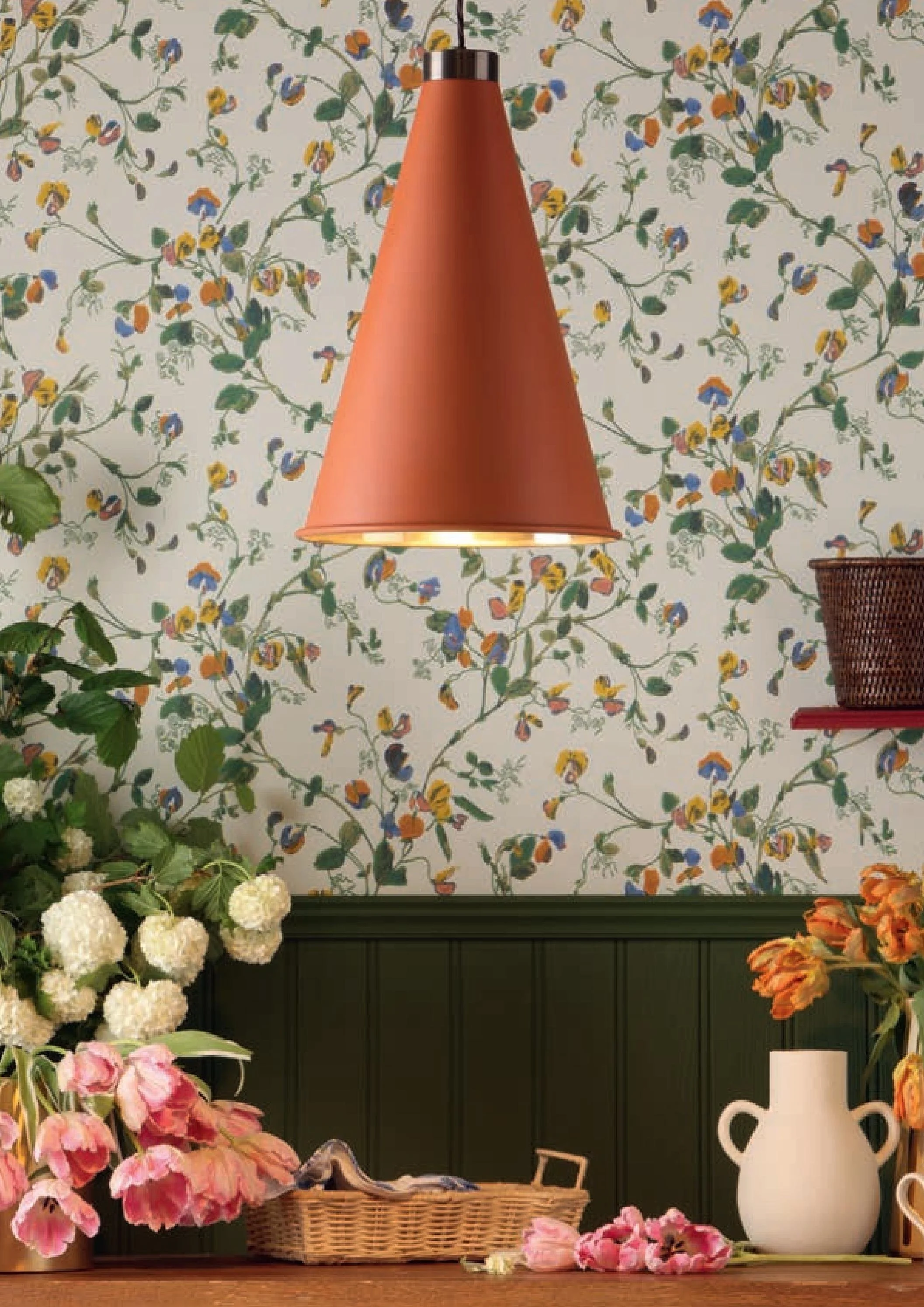 A wall with a colorful floral wallpaper, a hanging modern lampshade, and a table with various flowers, a basket, and a white vase.