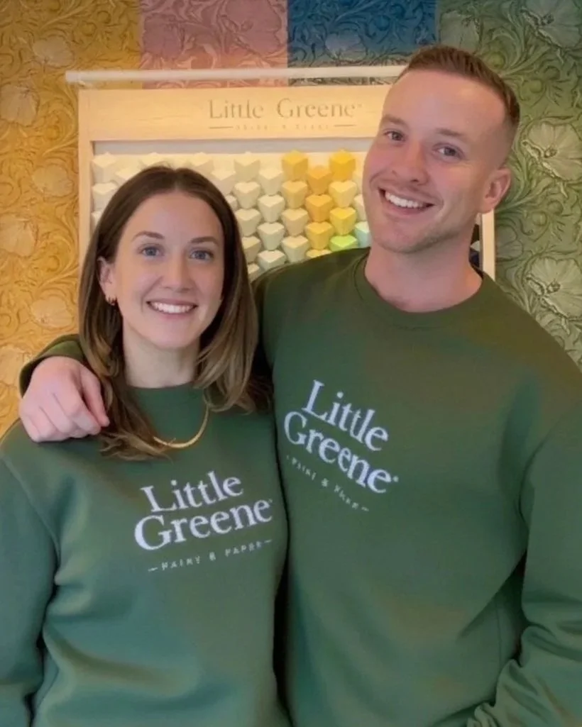 Two smiling people wearing matching green shirts with 'Little Greene' logo, standing in front of a colorful wallpaper and a decorative display of eggs.