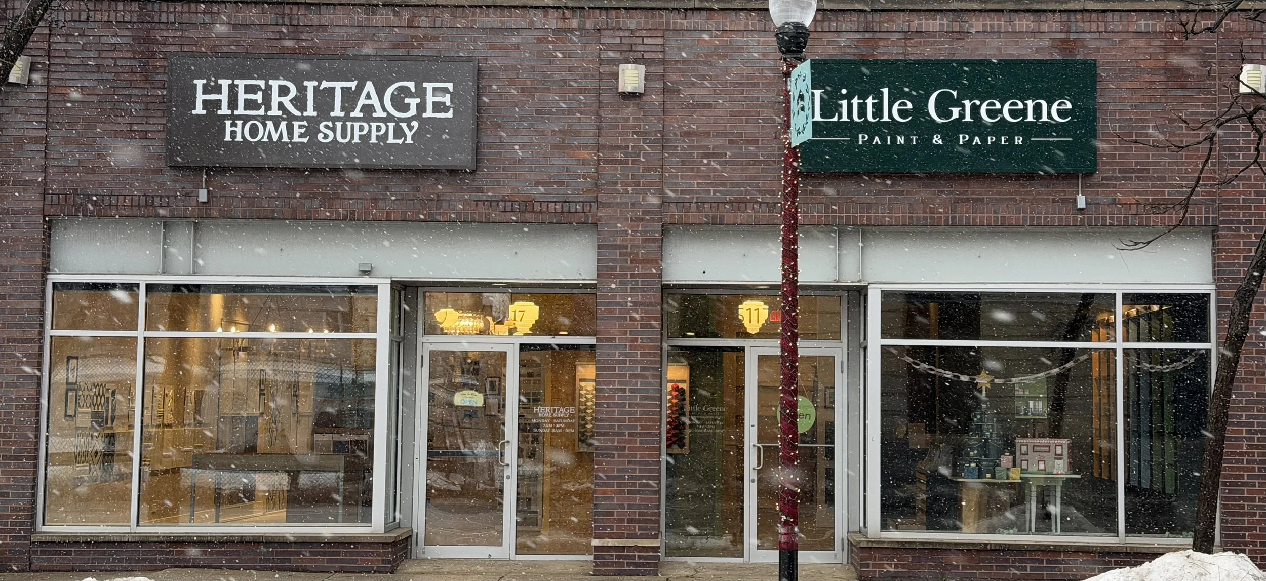 Brick storefront with signs for Heritage Home Supply and Little Greene Paint & Paper, large windows, snow falling, winter scene.