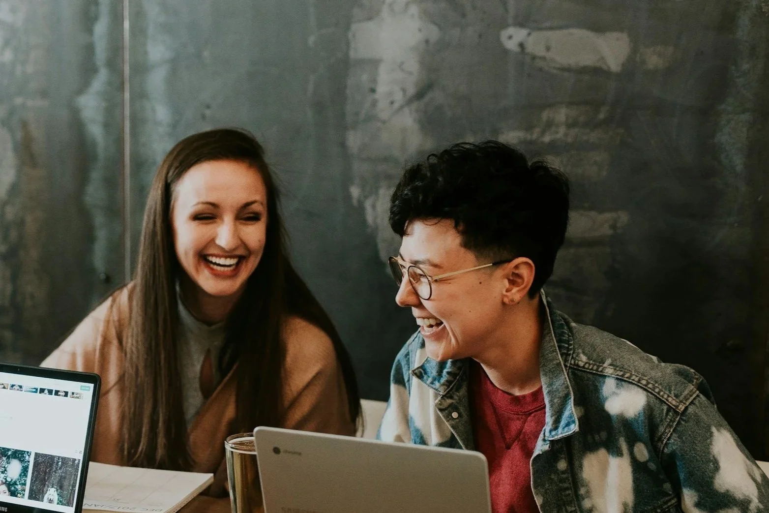 Two women are sitting at a table with laptops and notebooks, laughing together in a casual setting.