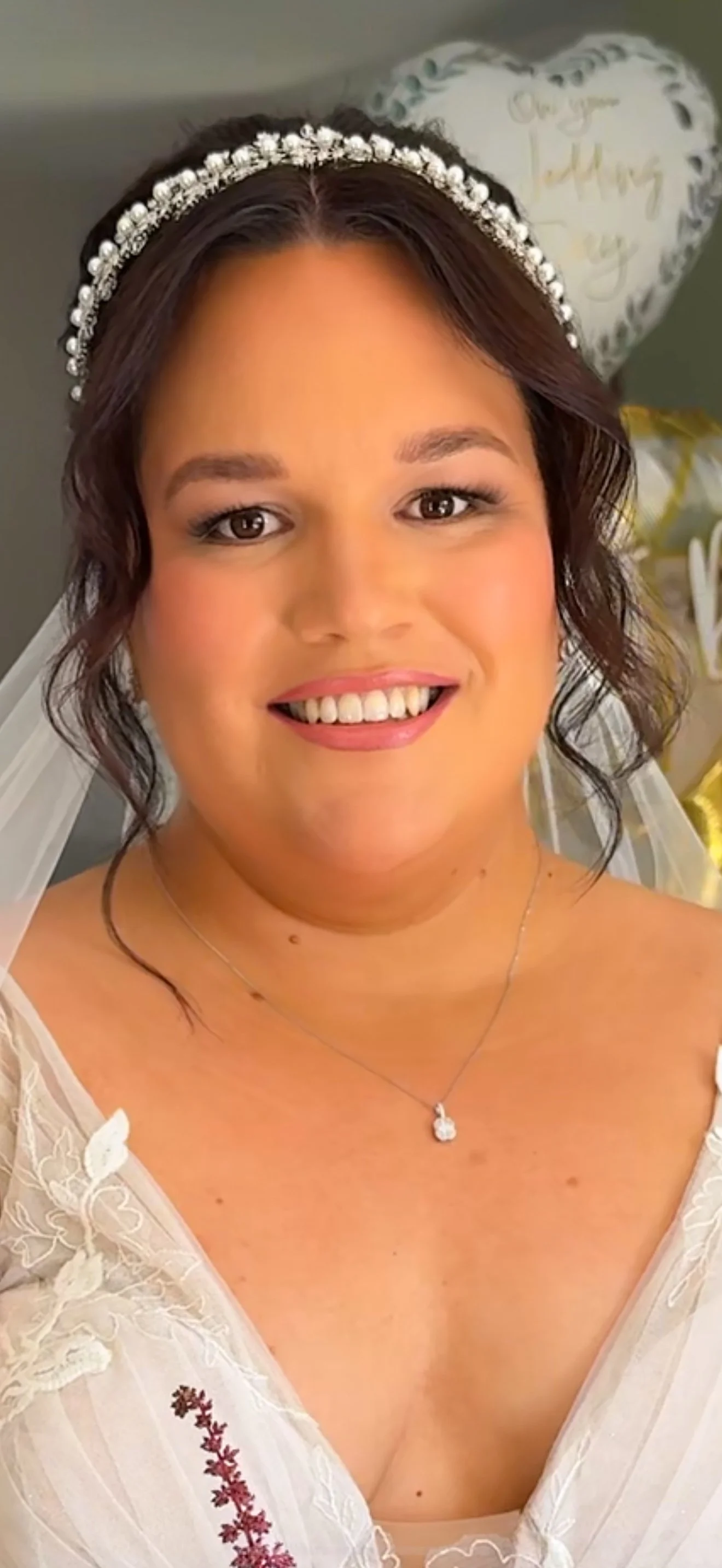 Close-up of a smiling bride in a wedding dress with a pearl headband, jewellery, and sheer fabric with floral embroidery.