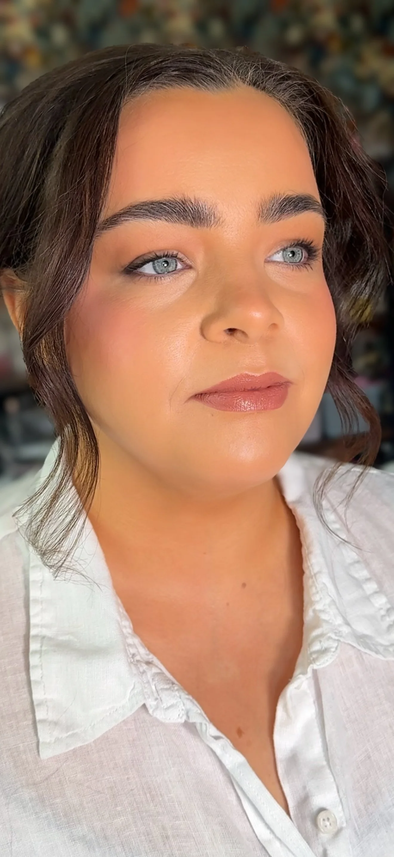 Close-up of a bride with blue eyes, dark brown hair styled in loose waves, wearing a white collared shirt, showing her makeup with a blurred background.