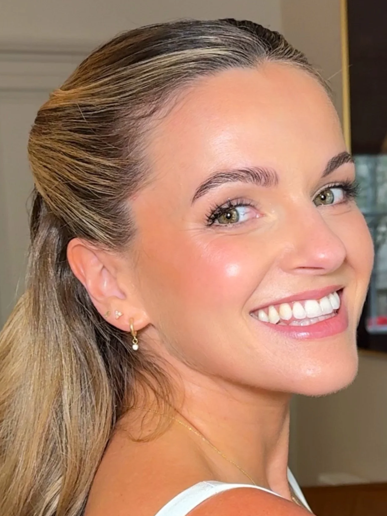 Close-up of a smiling bride with blonde hair, wearing earrings and a necklace, with a blurred background.