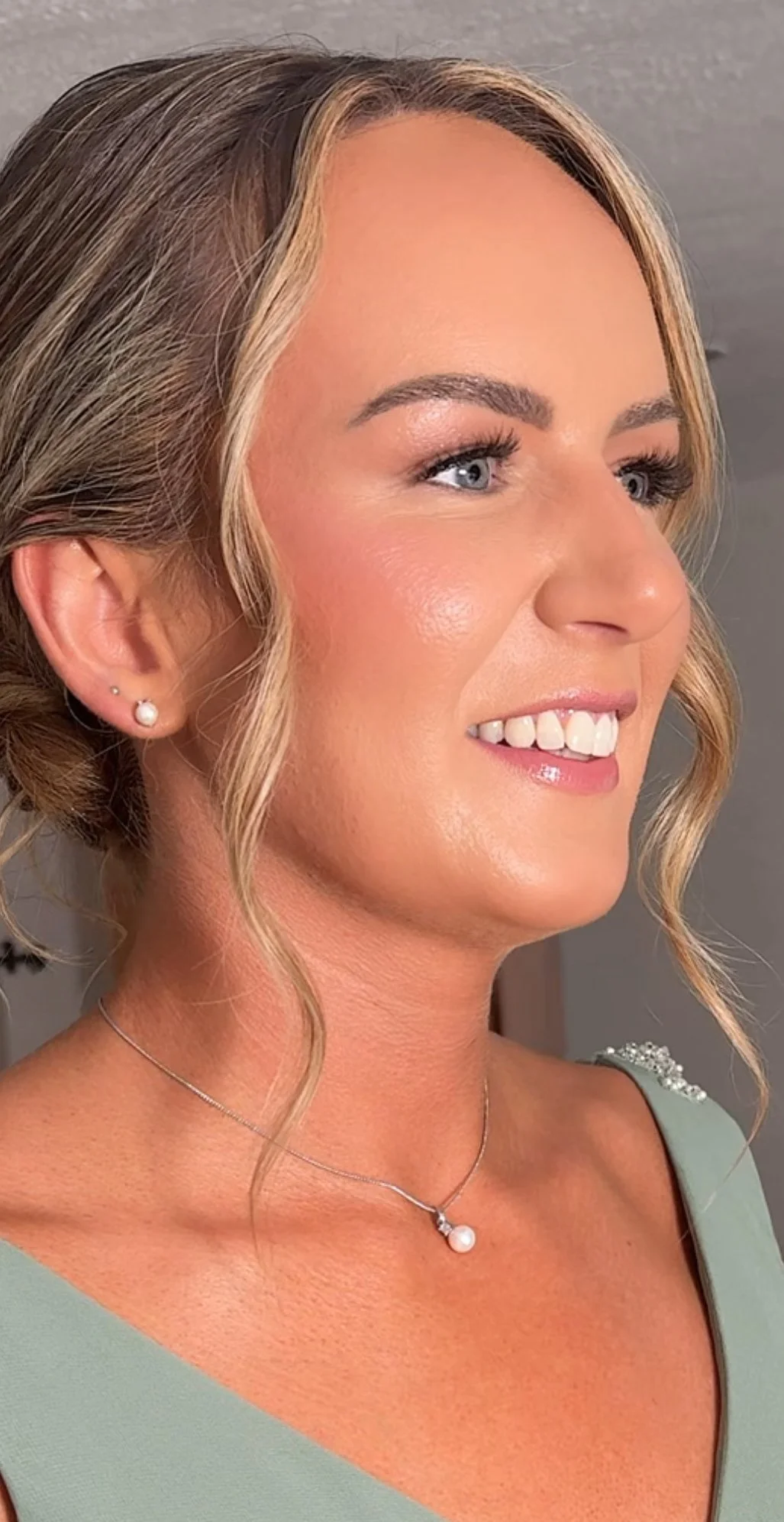 Close-up of a bridesmaid with blonde hair styled in loose waves, wearing pearl earrings and a pearl necklace, smiling slightly, in a light green dress with decorative shoulder detailing.