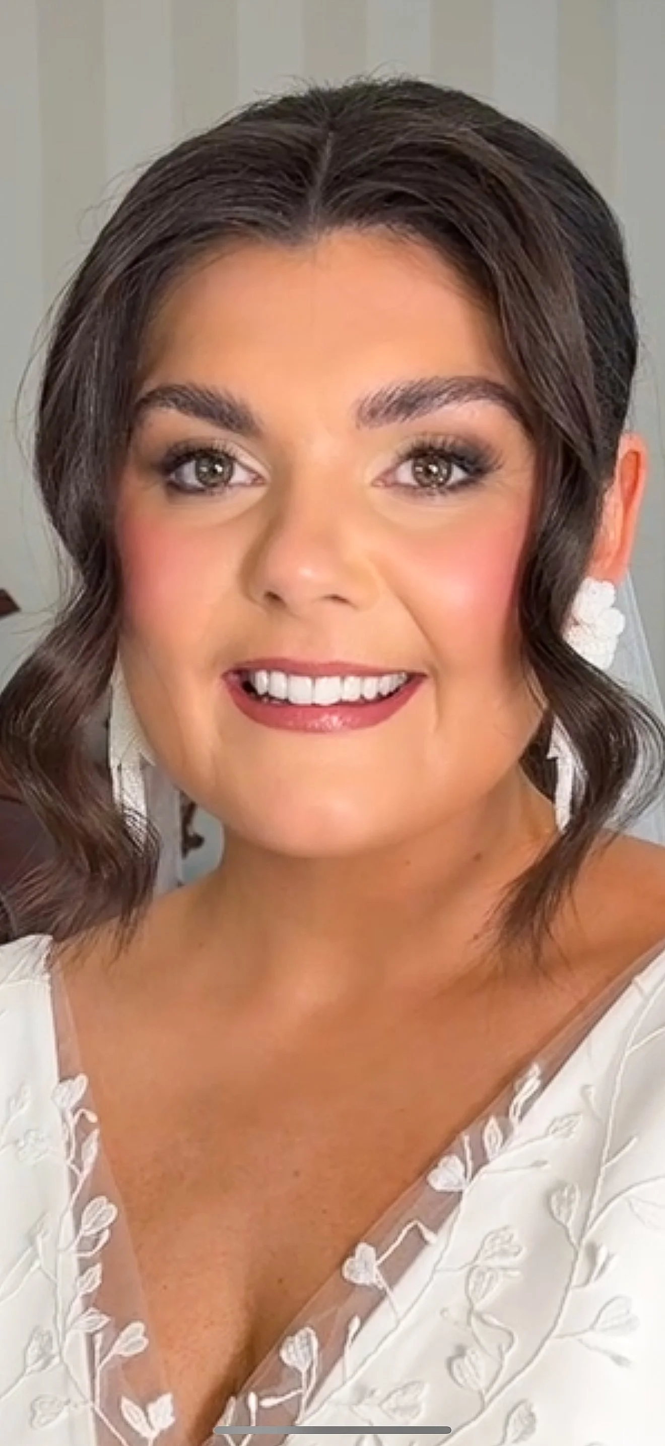 An Irish bride with shoulder-length wavy brown hair, smiling with beautiful, glowy makeup, wearing a white dress with floral embroidery and white earrings.