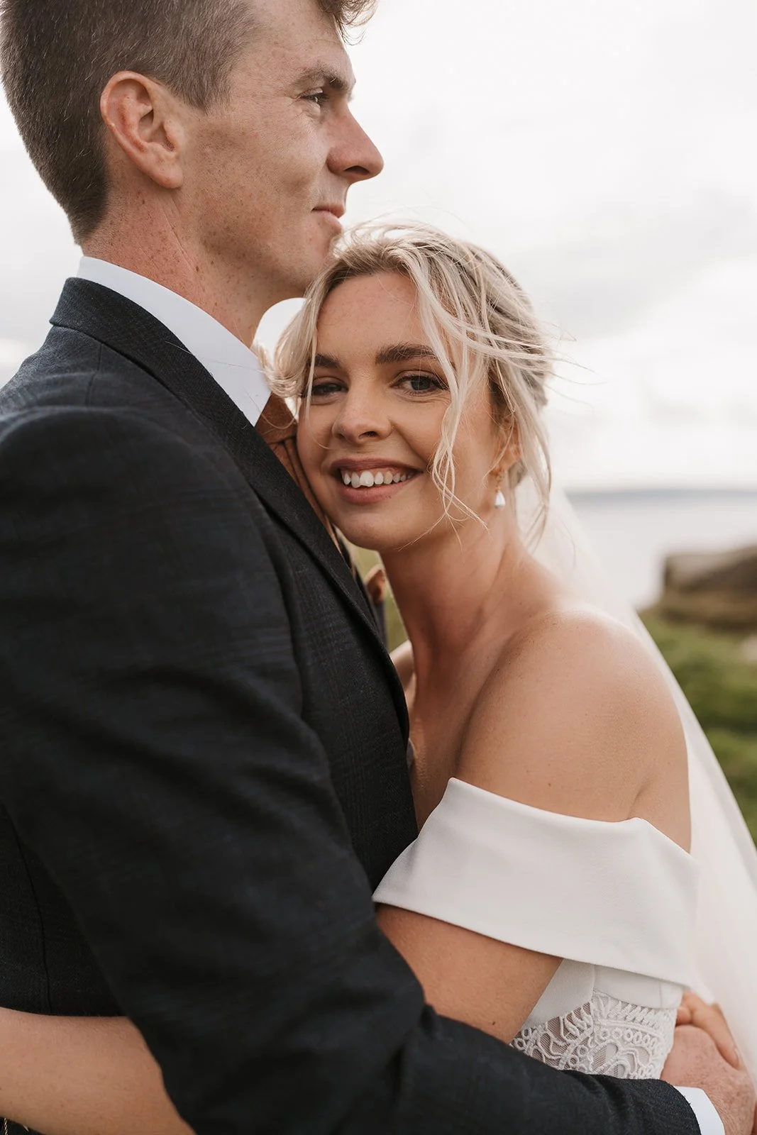 A smiling woman with blonde hair in an off-the-shoulder white dress embracing a man in a black suit outdoors.