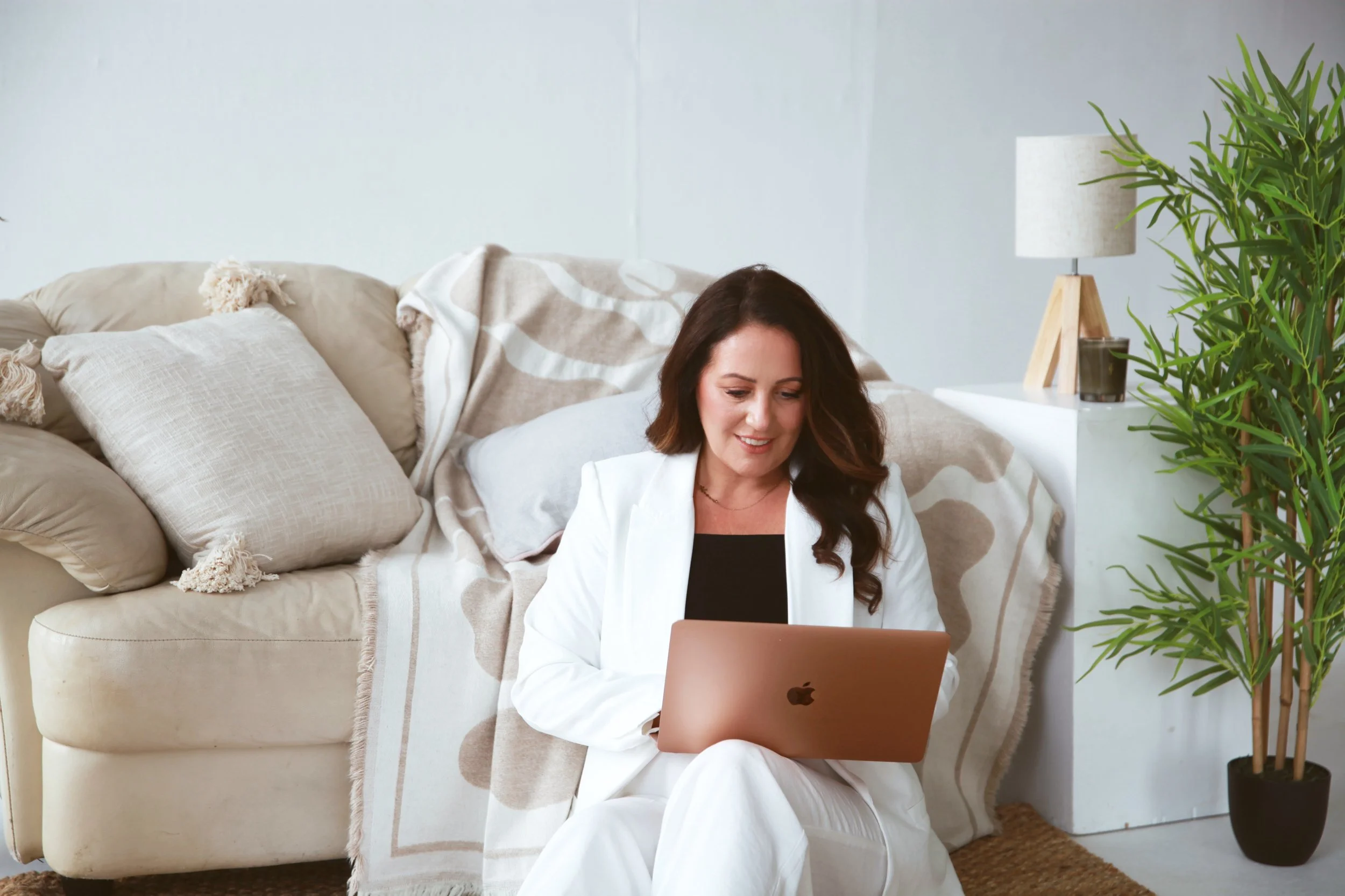 Bridal mentor sitting on the floor with a laptop, smiling, in a cozy living room with beige couch, pillows, blanket, plant, and side table with lamp.