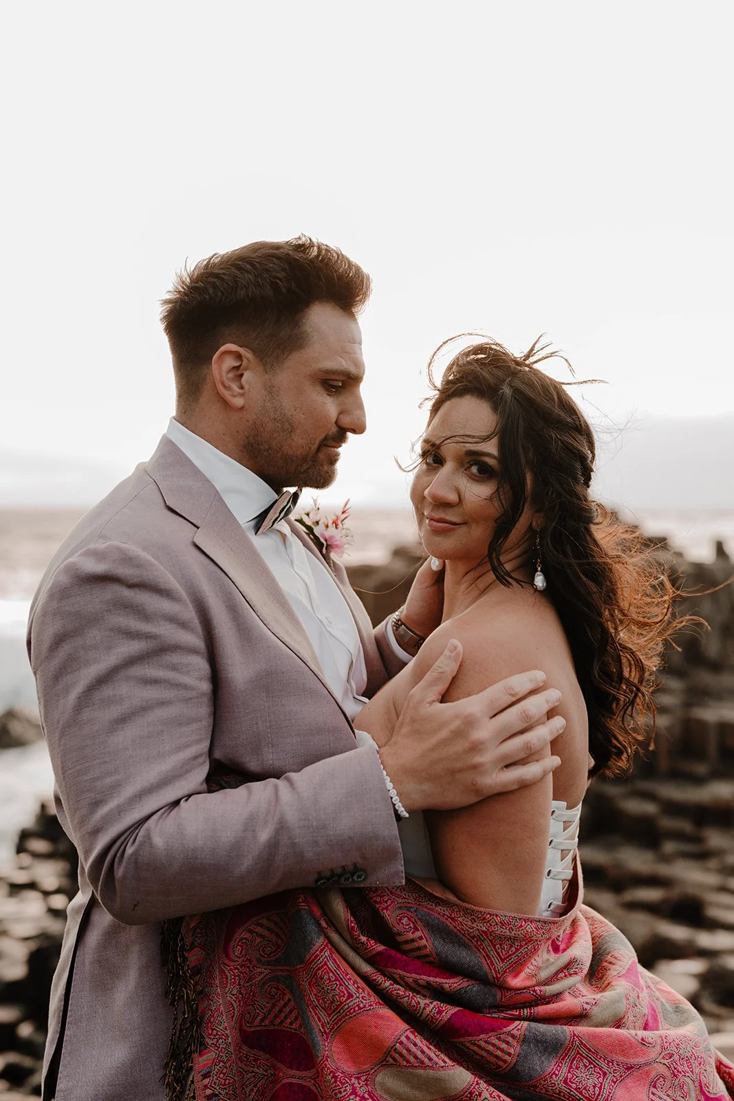 A couple holding each other affectionately on a rocky beach at sunset, dressed in formal attire with a pink and red patterned blanket draped over them.