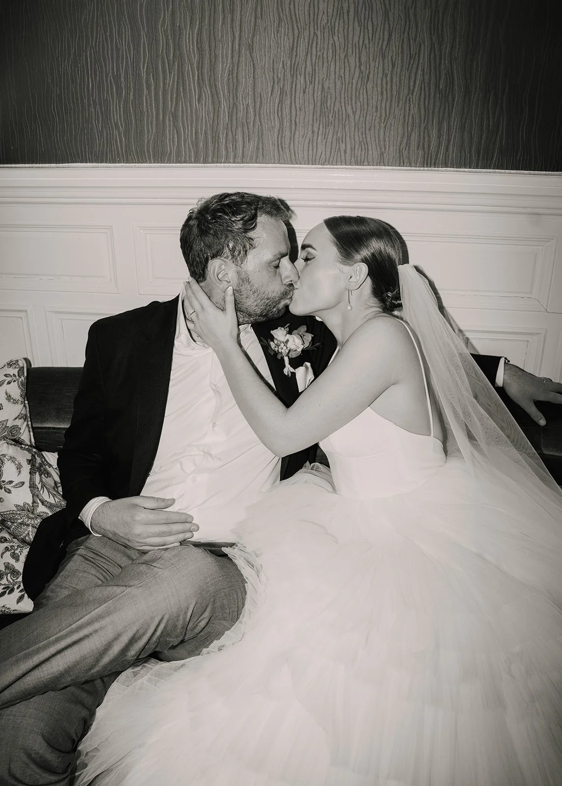 A black and white photo of a bride and groom sharing a kiss, seated closely on a couch in wedding attire, with the bride's hands gently on the groom's face.