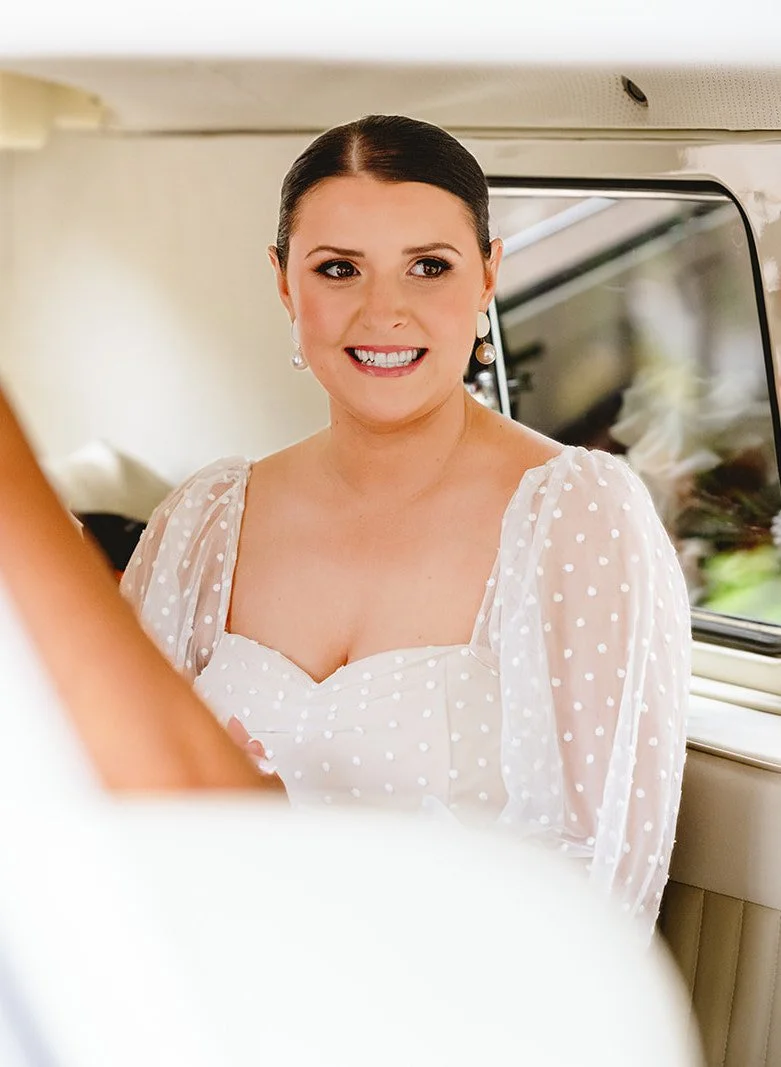 A smiling woman with dark hair in a bun, wearing pearl earrings and a white polka-dot dress with puffy sleeves, is inside a vehicle.