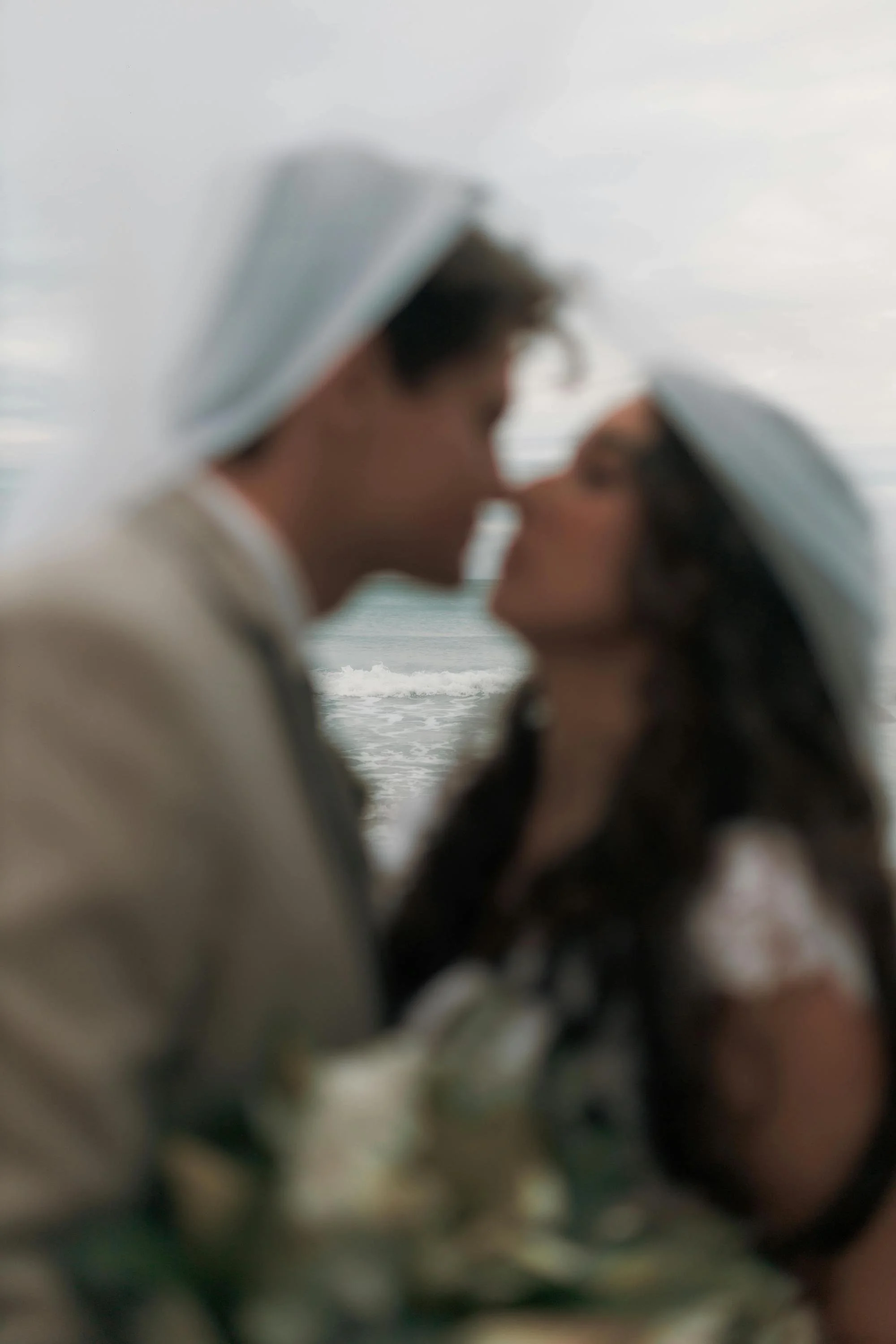Blurred image of a man and woman in wedding attire, wearing white hats, about to kiss with a beach and overcast sky in the background.