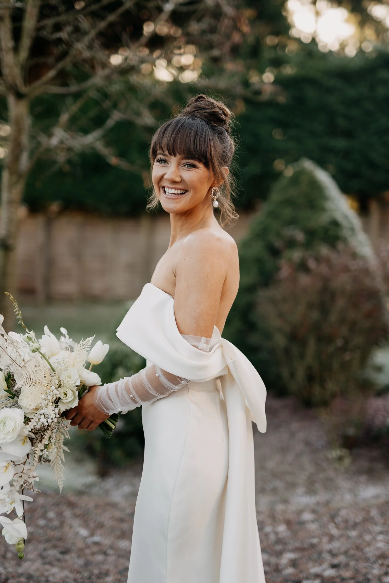 A smiling bride with dark hair styled in an updo with bangs, wearing a strapless white wedding gown with an off-the-shoulder bow and sheer long sleeves, holding a white floral bouquet outdoors.