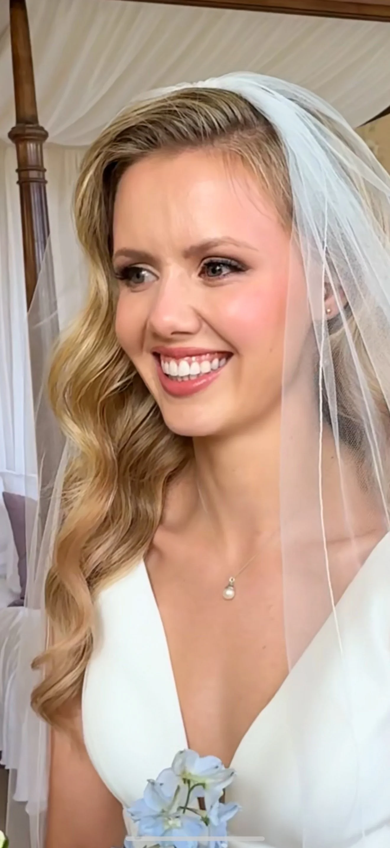 A smiling bride with long, wavy blonde hair wearing a white dress and veil, holding light blue flowers, on her wedding day.