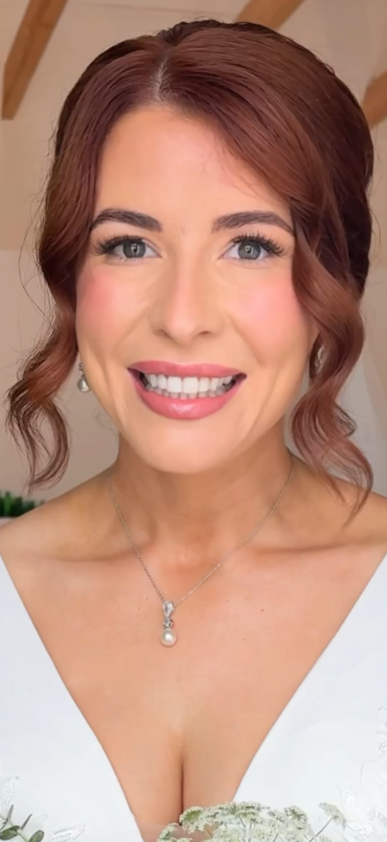 Close-up of a smiling bridewith reddish-brown wavy hair, wearing silver jewellery and a white wedding dress, indoors with a beige and wooden background.