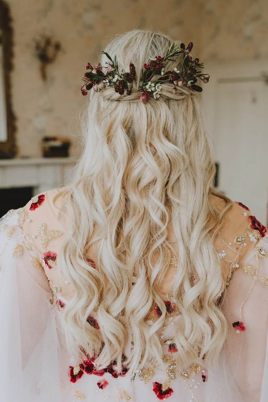 Back of a woman with long, wavy blonde hair wearing a floral crown and a dress decorated with red, gold, and pearl embellishments.