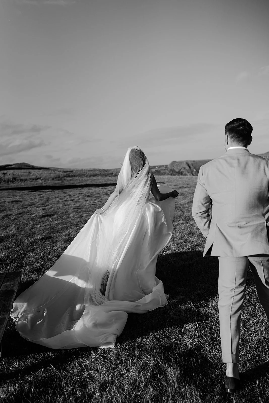 Black and white photo of a bride in a wedding gown and a groom in a suit walking outside on a grassy field, with distant hills and sky in the background.