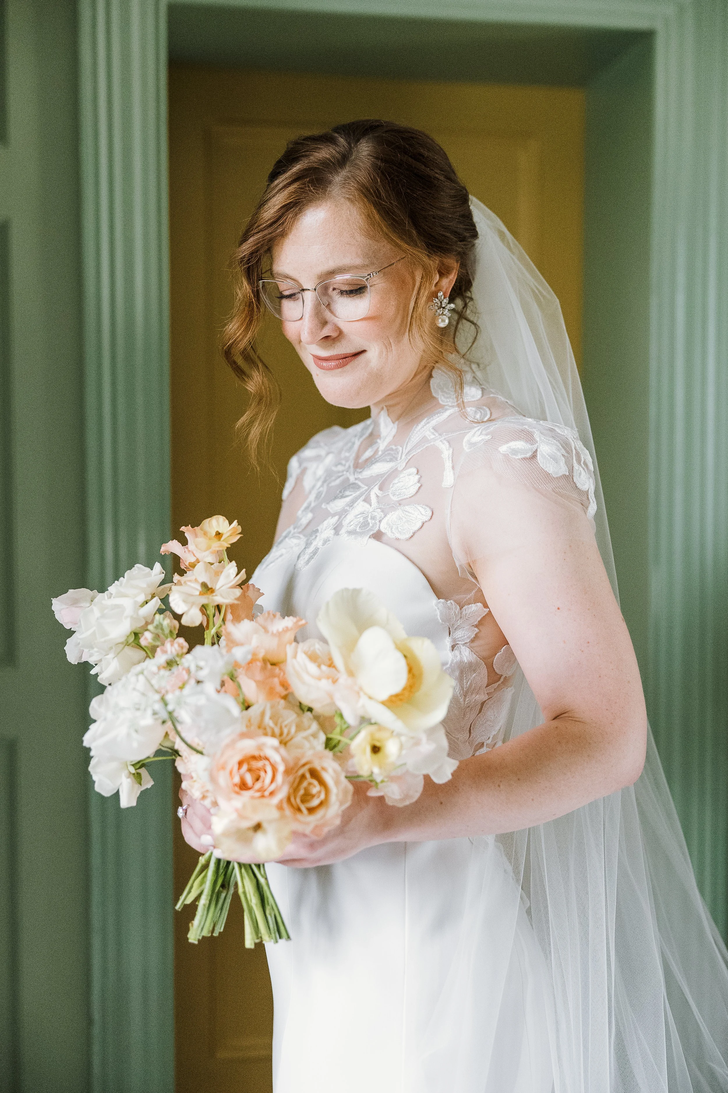 A bride with shoulder-length wavy hair, wearing glasses, earrings, a white wedding dress with lace details, and a veil, holding a bouquet of light-colored flowers, standing indoors near a green door frame.