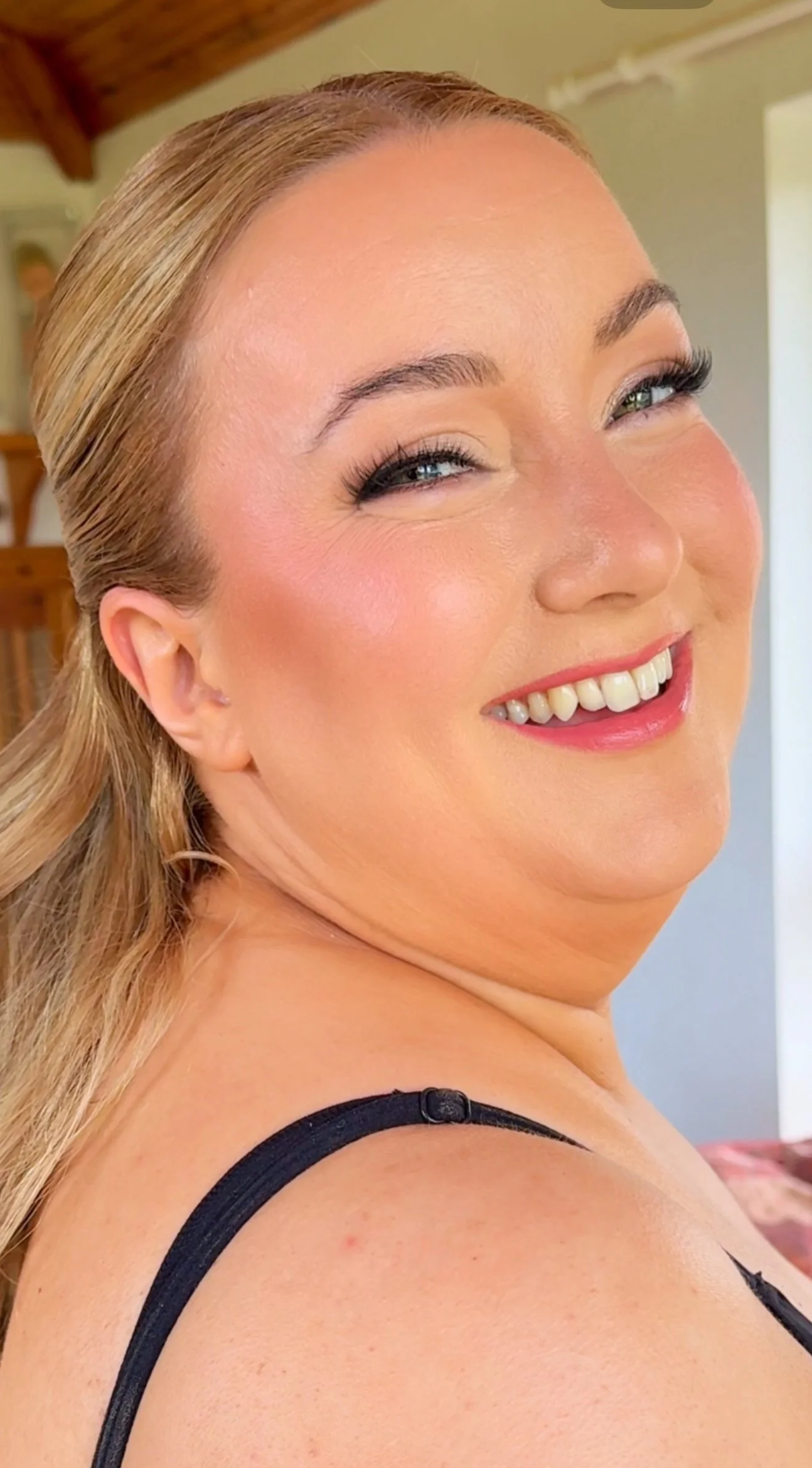 Close-up of smiling elopement bride with blonde hair, wearing makeup, with a black strap on her shoulder, and a background with a wooden ceiling and wall.