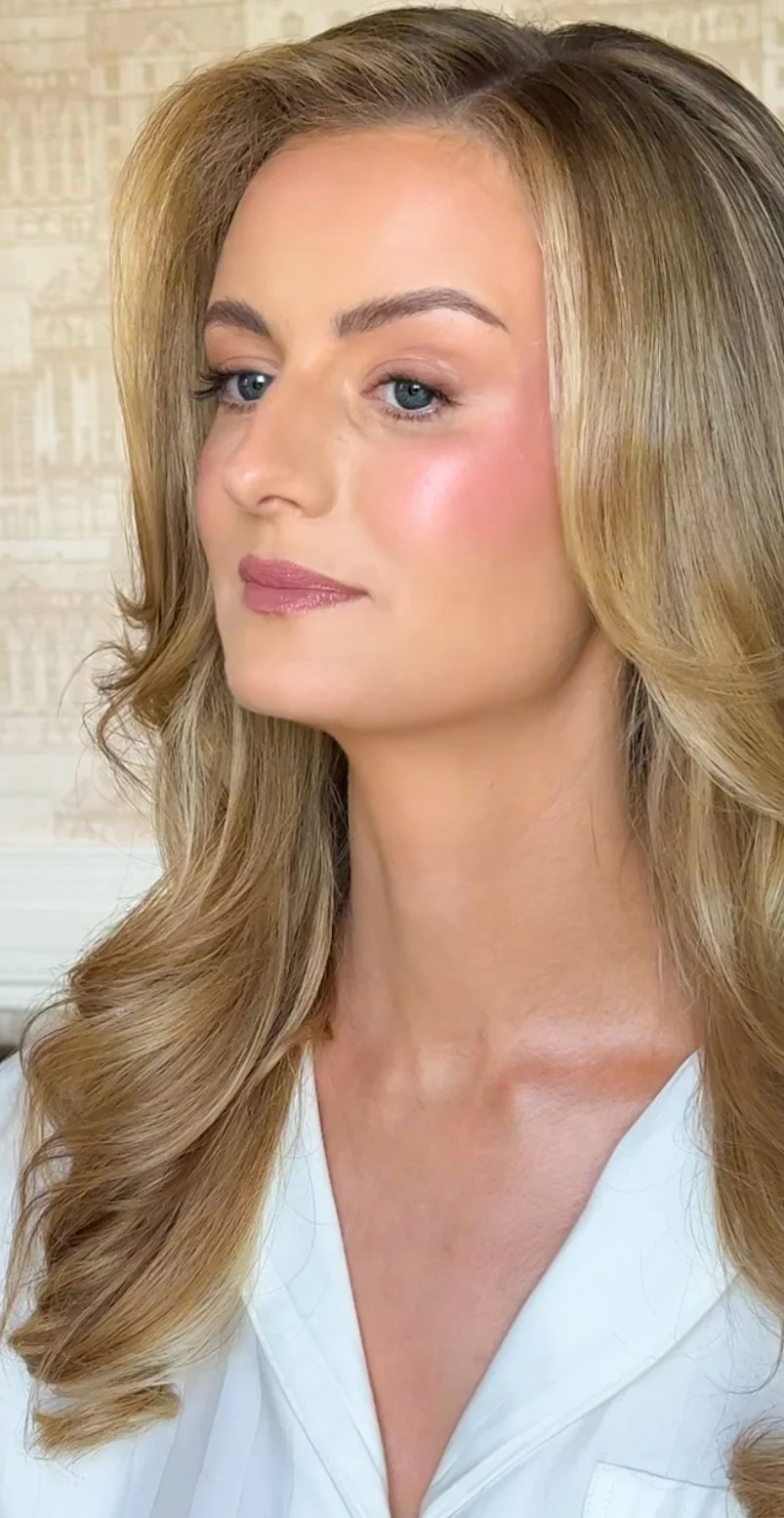 Close-up of a bride with long blonde hair, blue eyes, wearing light makeup and a white top, against a background of wallpaper with architectural sketches.