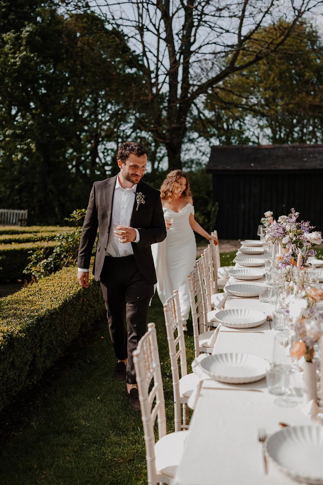 A man and a woman walking beside an outdoor dining table decorated with flowers and tableware, set in a garden with trees.
