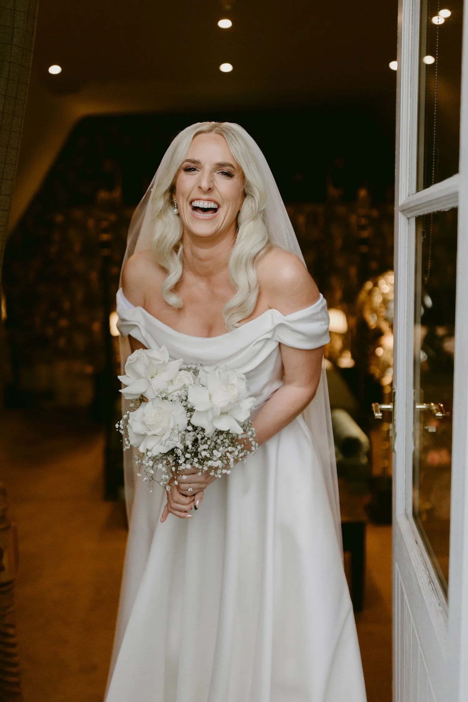 A bride with long blonde hair, wearing an off-shoulder white wedding dress and holding a bouquet of white roses and baby's breath, standing indoors with warm lighting at night.