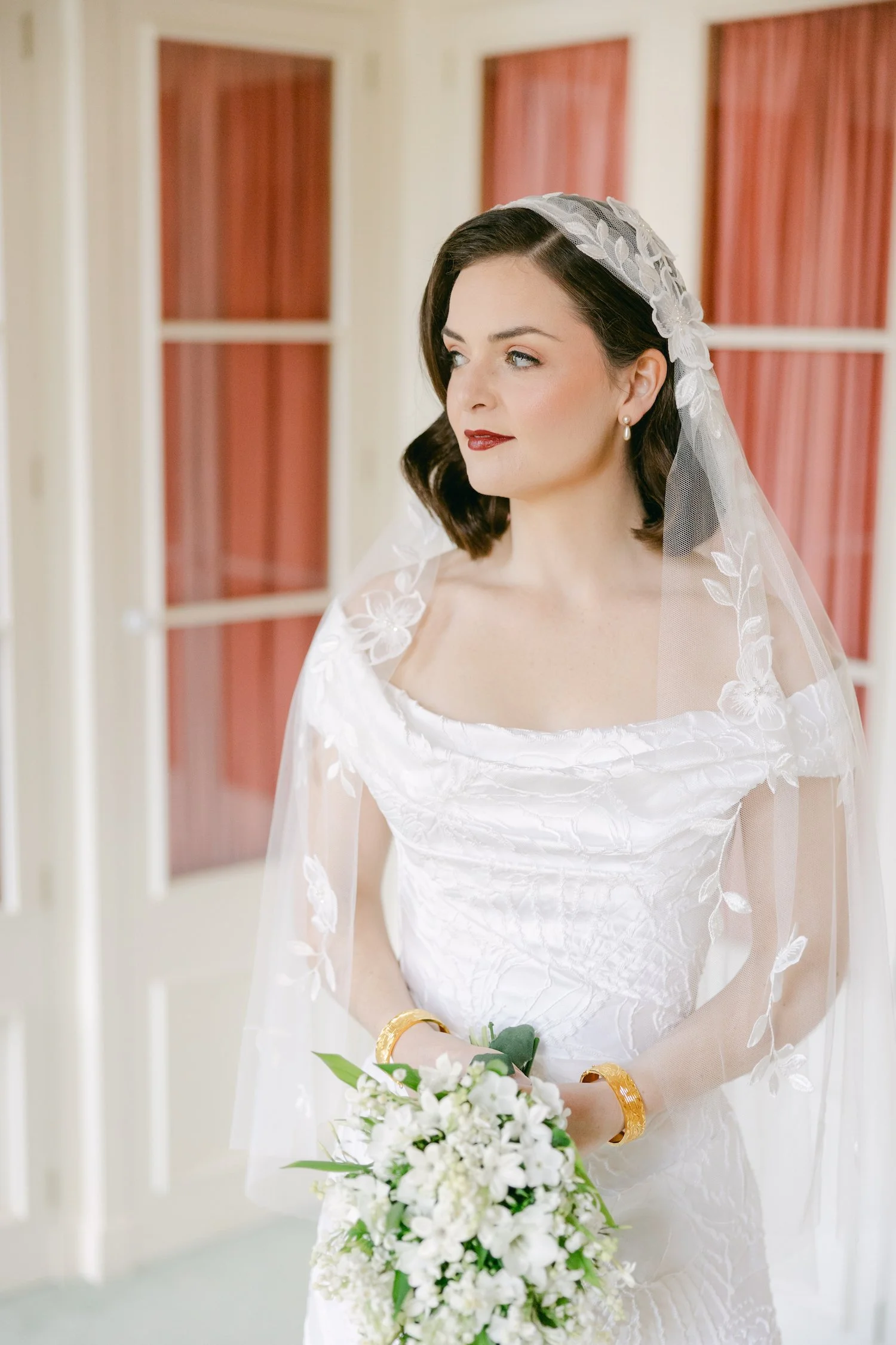 A woman in a wedding dress holding a bouquet of white flowers, with dark hair, red lipstick, and a lace veil, standing indoors with windows in the background.