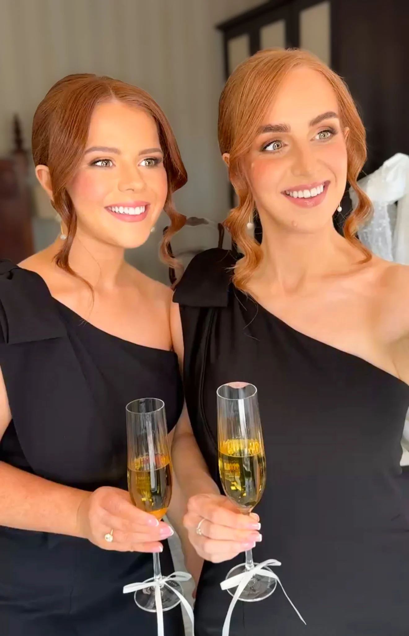 Two bridesmaids with red hair smiling and holding glasses of champagne, dressed in black dresses, in a well-lit room.