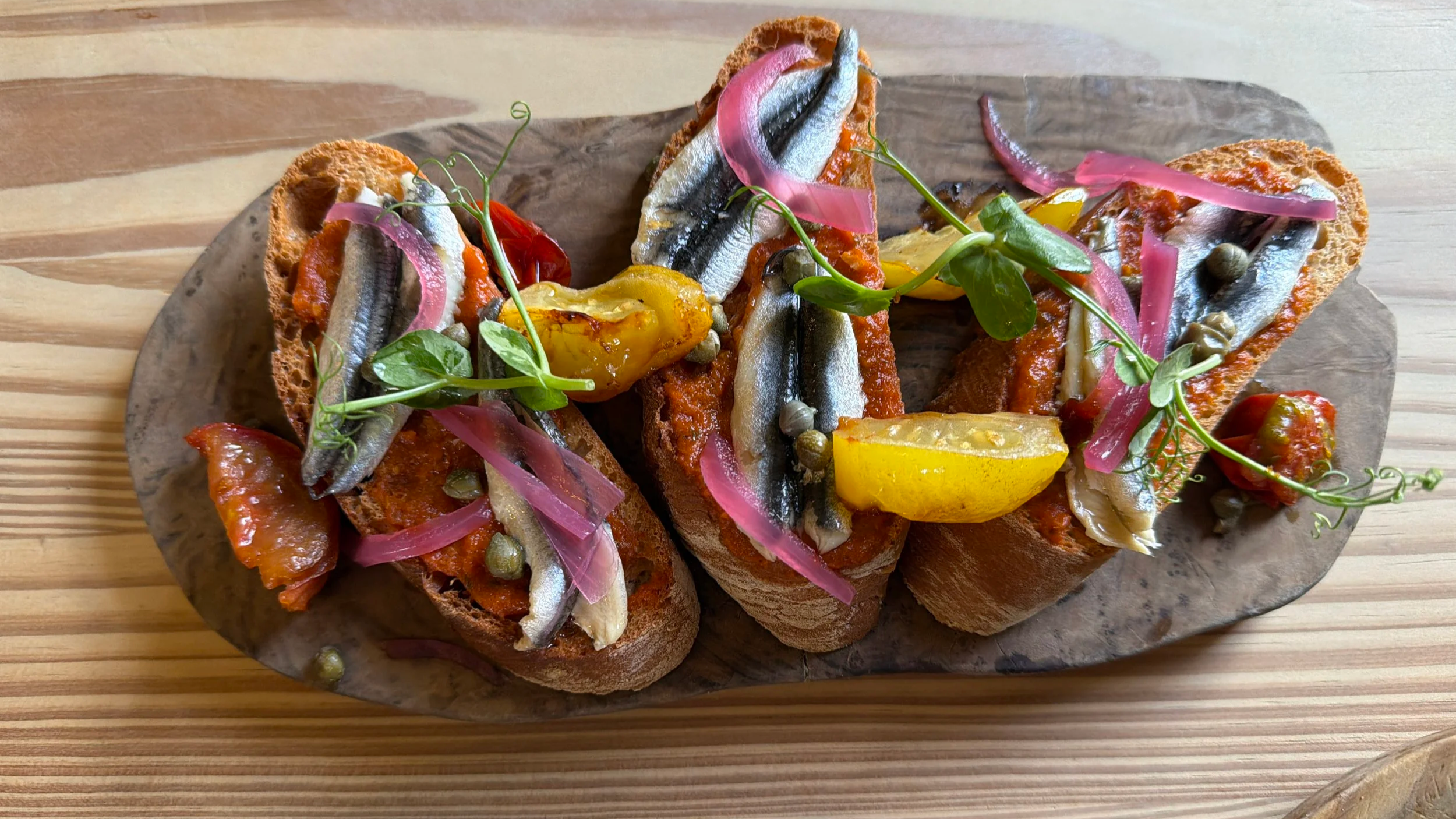 Open-faced sandwiches topped with anchovies, pickles, capers, pickled onions, and fresh herbs on toasted bread served on a wooden board.