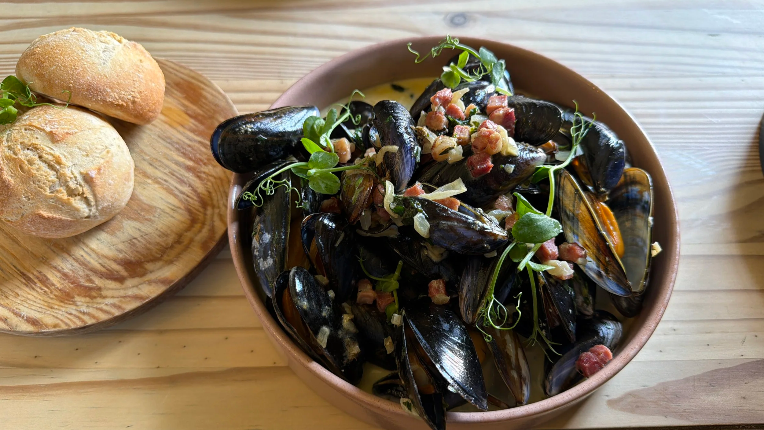 A bowl of steamed mussels garnished with herbs and bacon bits, served with bread rolls on a wooden tray.