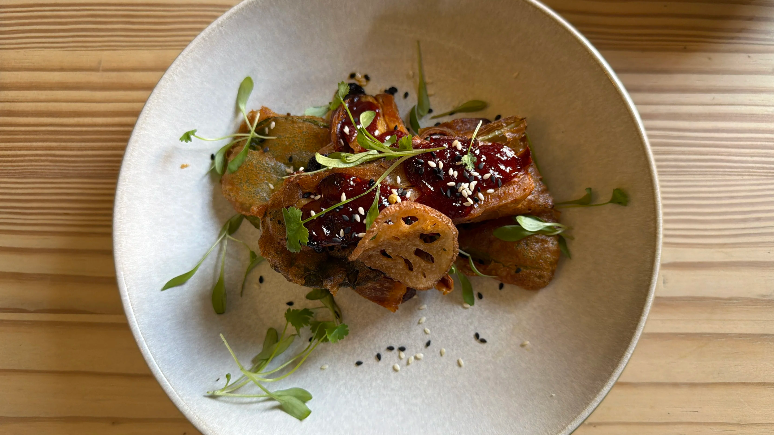 A dish of Asian-style fried tofu garnished with microgreens, black sesame seeds, white sesame seeds, and a dark sauce on top, served in a white bowl on a bamboo mat.