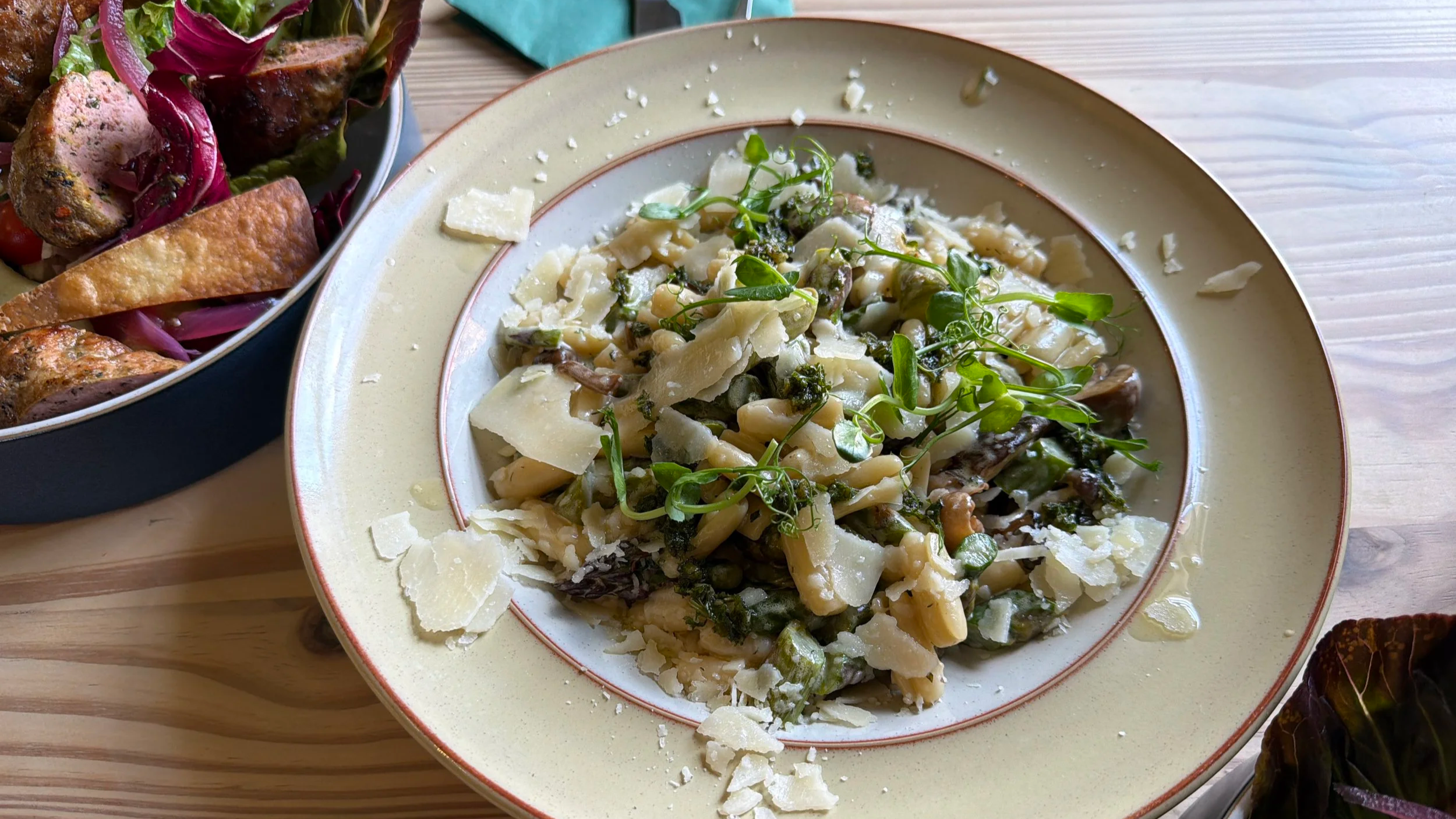 A plate of pasta with greens, grated cheese, and microgreens on top, next to a bowl of salad with roasted vegetables on a wooden table.