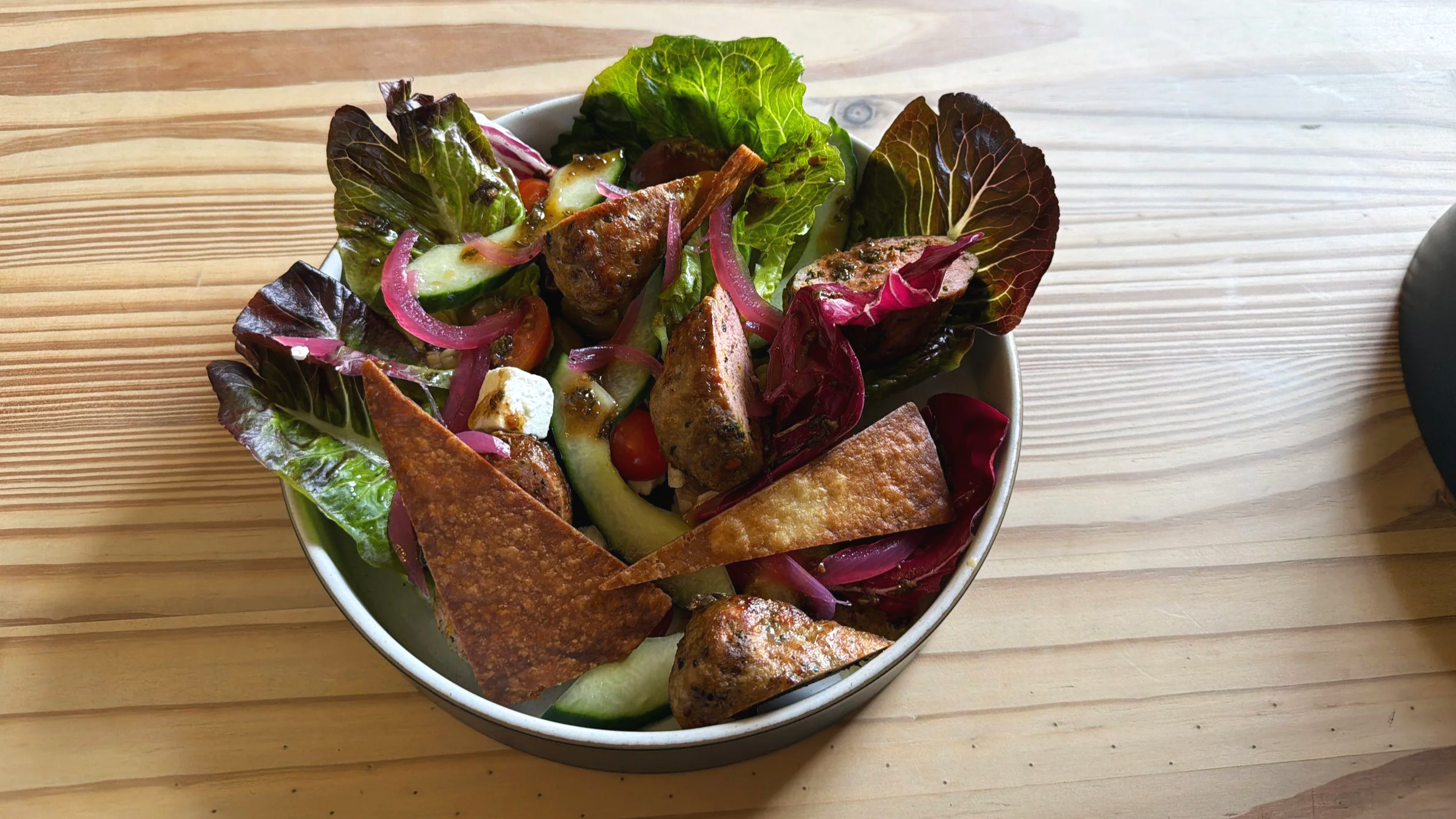 Salad in a white bowl with mixed greens, cherry tomatoes, cucumber, red onions, grilled chicken pieces, and crispy tortilla chips, on a wooden table.