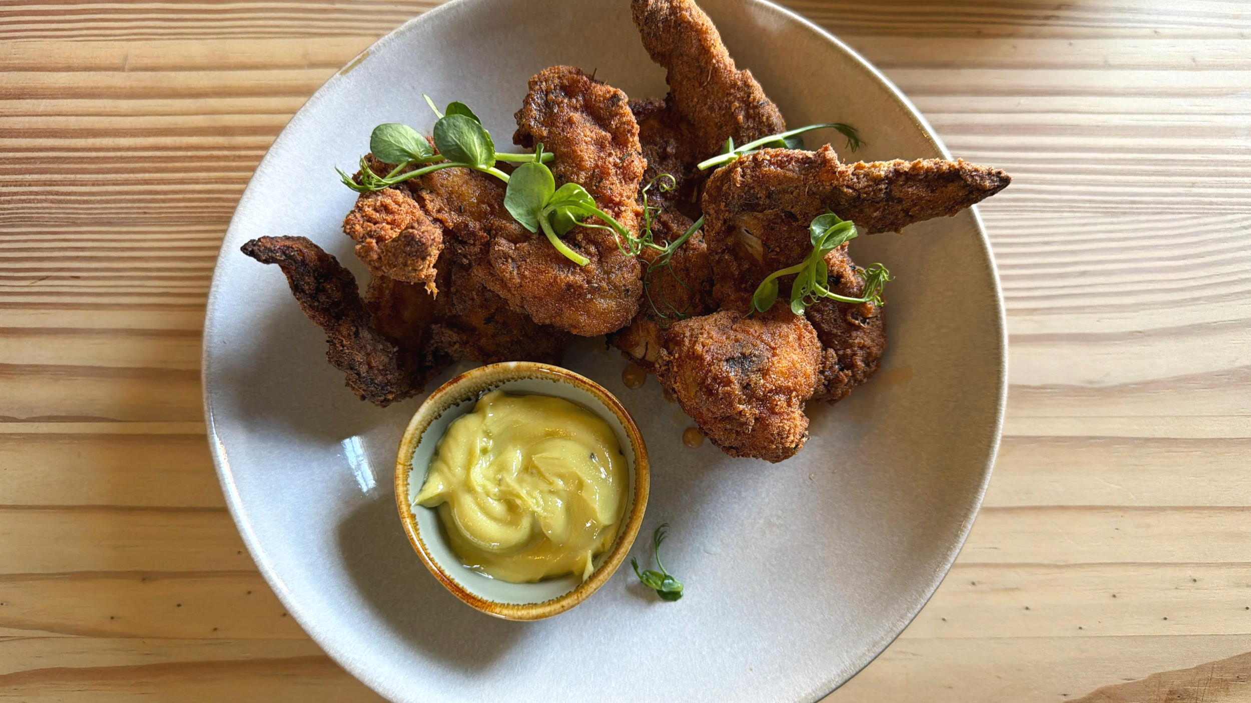 Fried chicken wings garnished with microgreens on a white plate, served with a small bowl of mustard sauce.