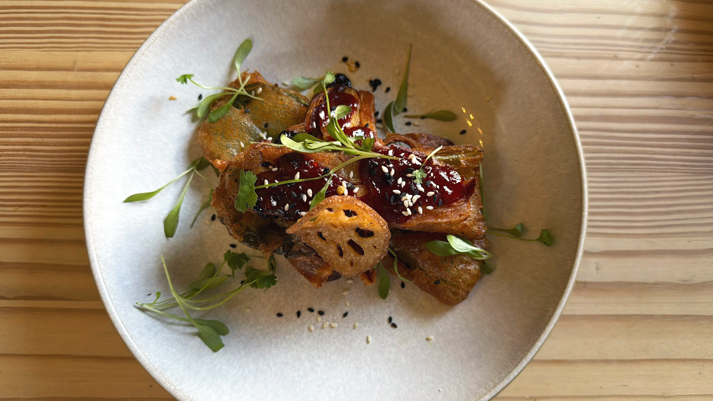 A dish with fried plantains, topped with berry sauce, sesame seeds, and garnished with microgreens on a white plate.
