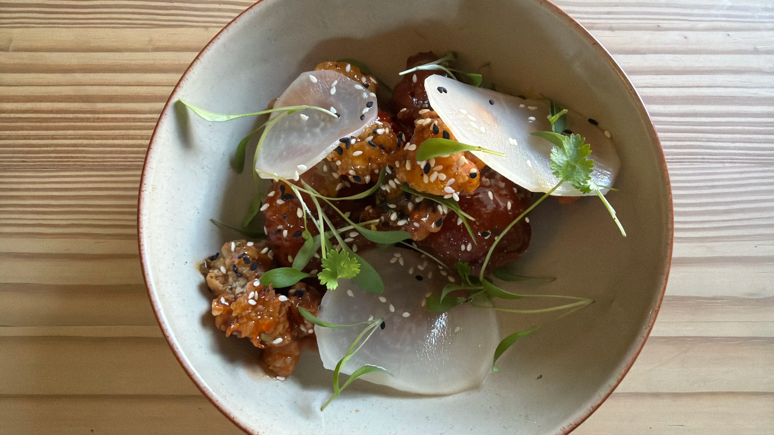 A bowl of Asian-style cuisine with spicy glazed chicken bites topped with sesame seeds, thinly sliced pickled radish, microgreens, cilantro, and sunflower seeds, served on a wood surface.