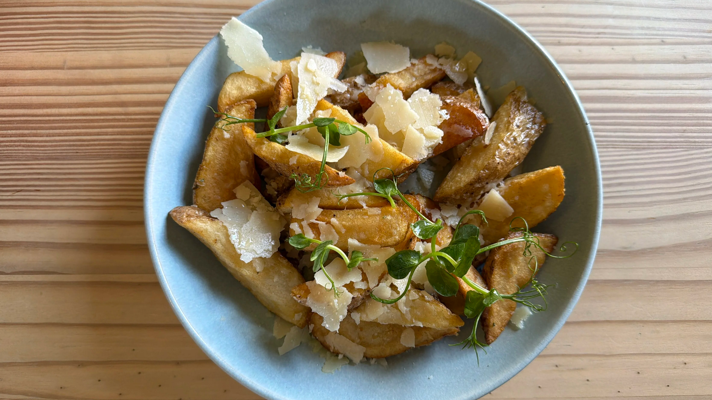 Bowl of roasted potato wedges topped with shaved cheese and garnished with fresh herbs on a wooden surface.