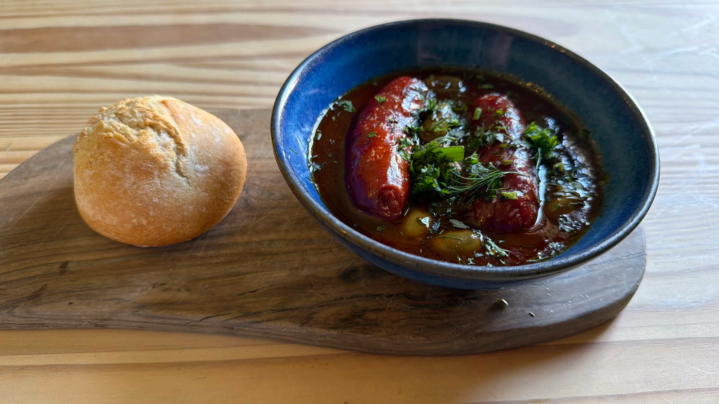 A bowl of moroccan-style meatball stew garnished with chopped parsley, next to a round bread roll on a wooden serving board.