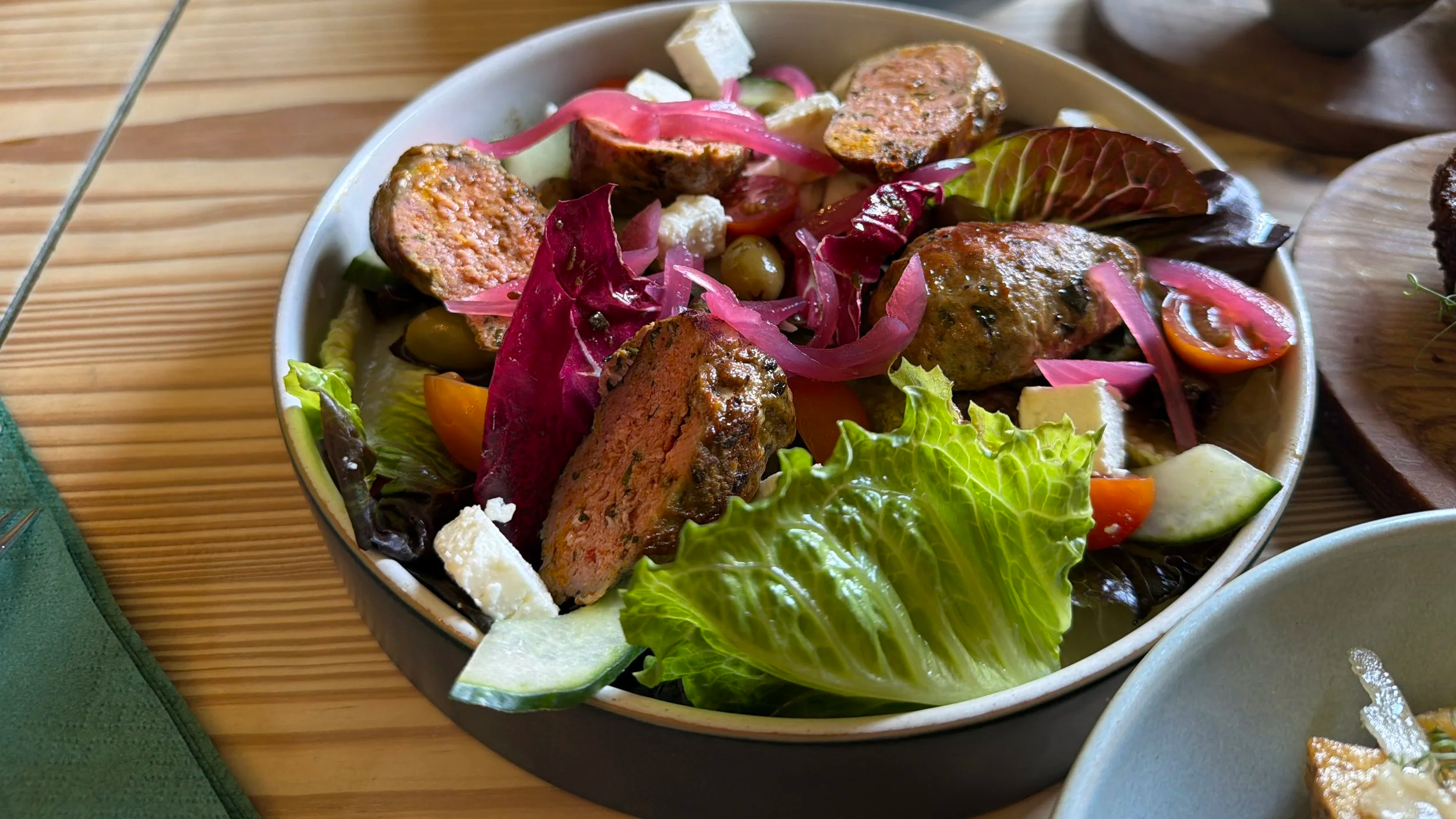 A bowl of fresh salad with mixed greens, cherry tomatoes, cucumbers, feta cheese, pickled red onions, grilled sausage slices, and green olives on a wooden table.